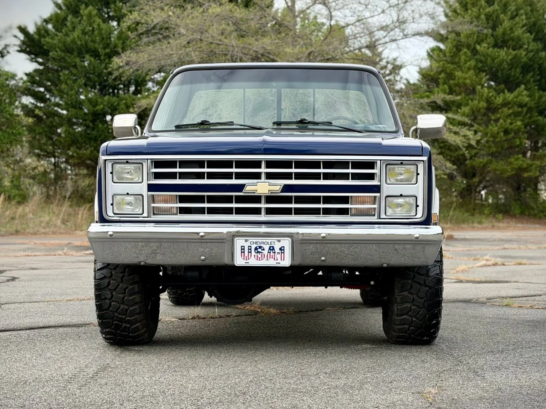 Front view of a vintage Chevrolet SUV with large tires, blue body, and a U.S.A. license plate, parked on an empty lot with trees in the background.