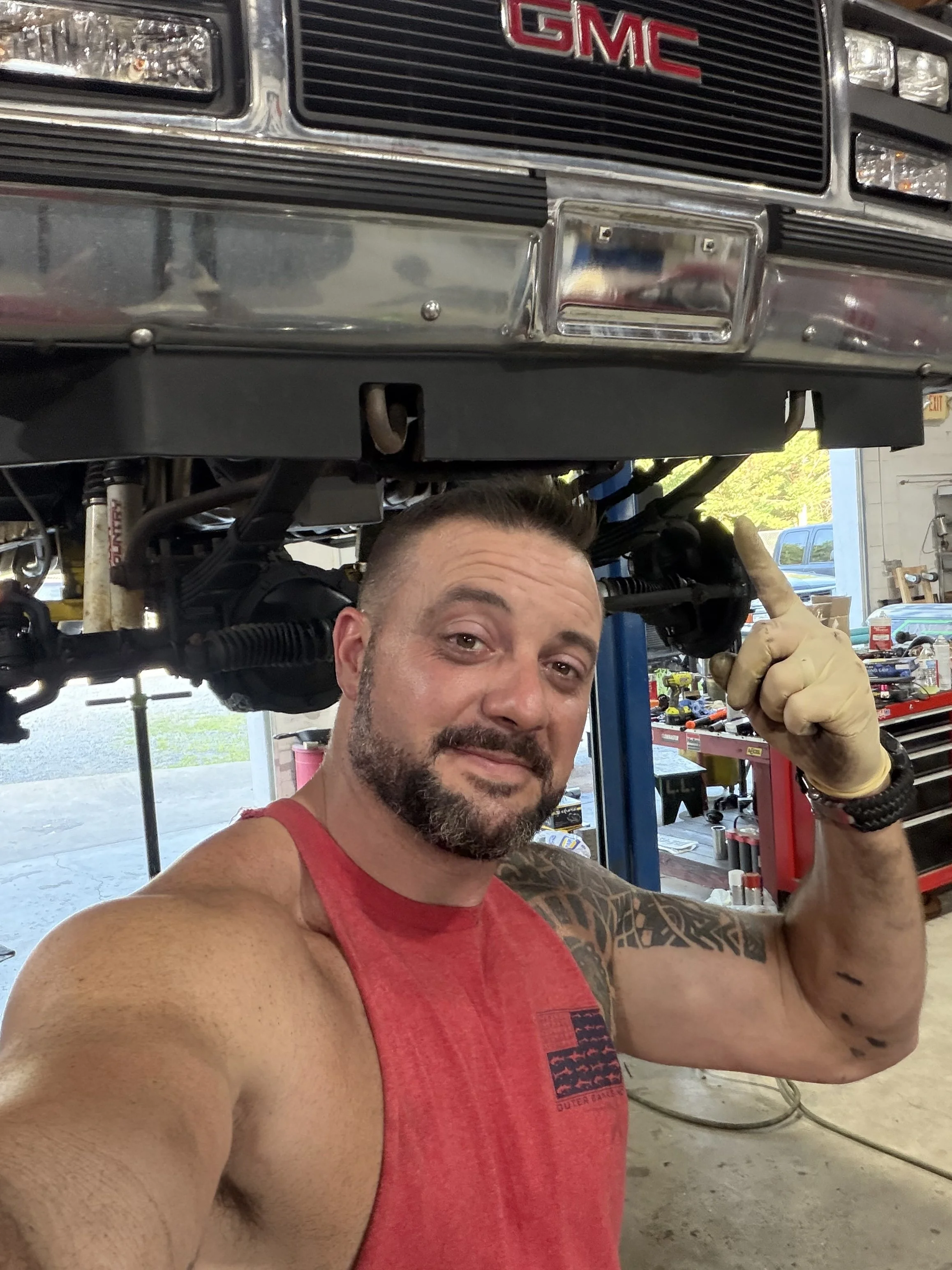 A man in a red sleeveless shirt taking a selfie in a workshop, with a vehicle lifted on a hydraulic lift visible above him.