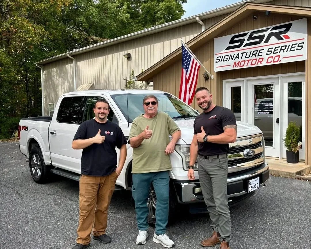 Three men standing in front of a white Ford truck outside a building with a sign that reads 'SSR Signature Series Motor Co.' and an American flag. The men are smiling, with two giving thumbs-up gestures.