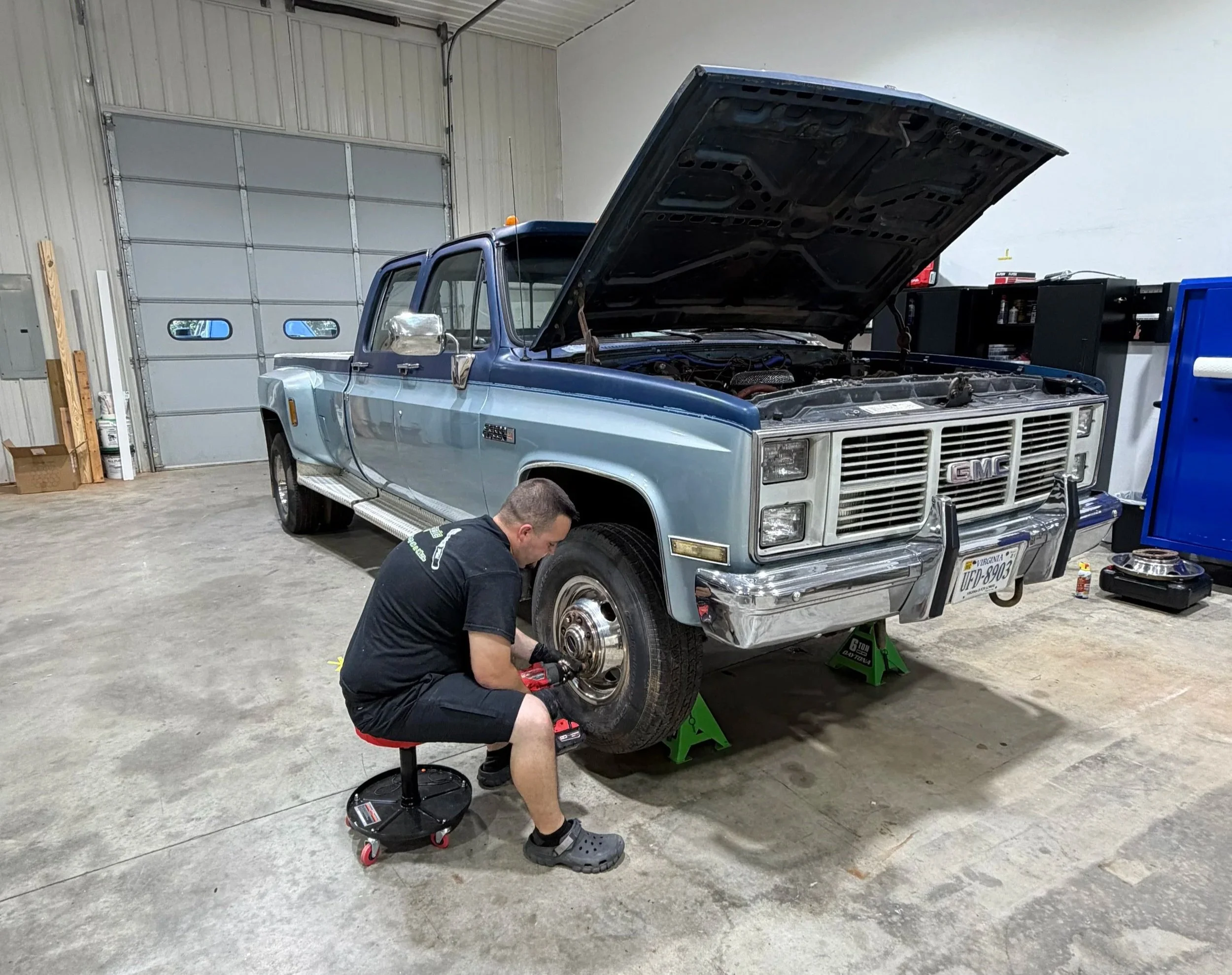 A person working on a blue GMC pickup truck inside a garage. The hood of the truck is open, and the person is kneeling while using tools. The garage has a large gray door, shelves, and various tools and equipment around.