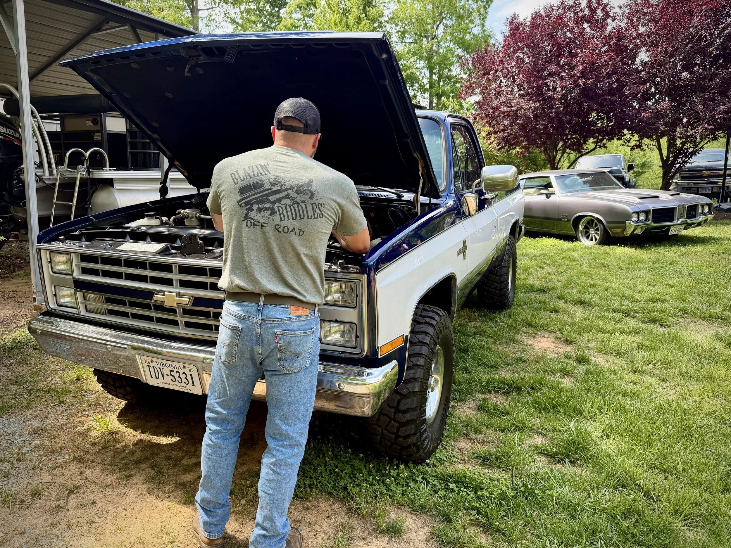 Chris Stiltner of Signature Series Motor Company with a black cap and gray T-shirt working on the engine of a vintage Chevrolet pickup truck with the hood open, parked on a grassy area with trees and other cars in the background.