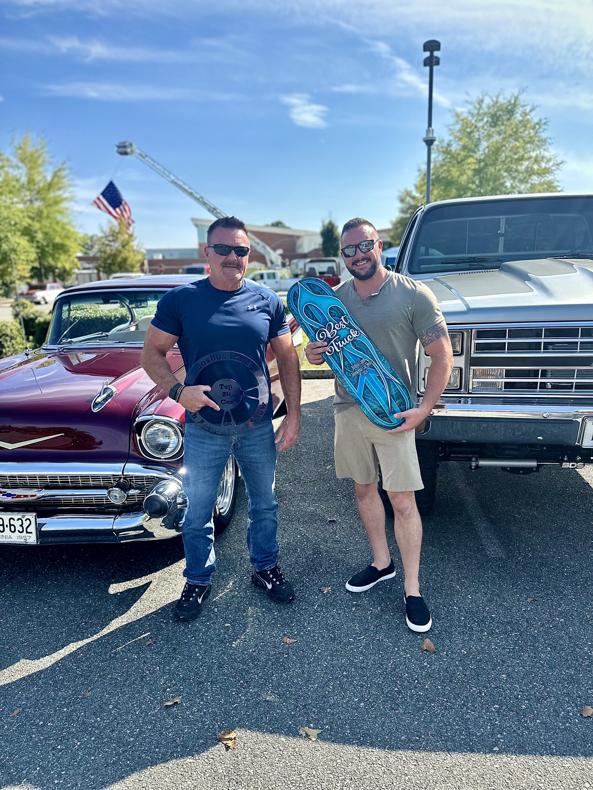 Two men, Chris Stiltner of Signature Series Motor Company and his father, standing in a parking lot in front of vintage cars, holding awards, with a fire truck ladder and American flag visible in the background.