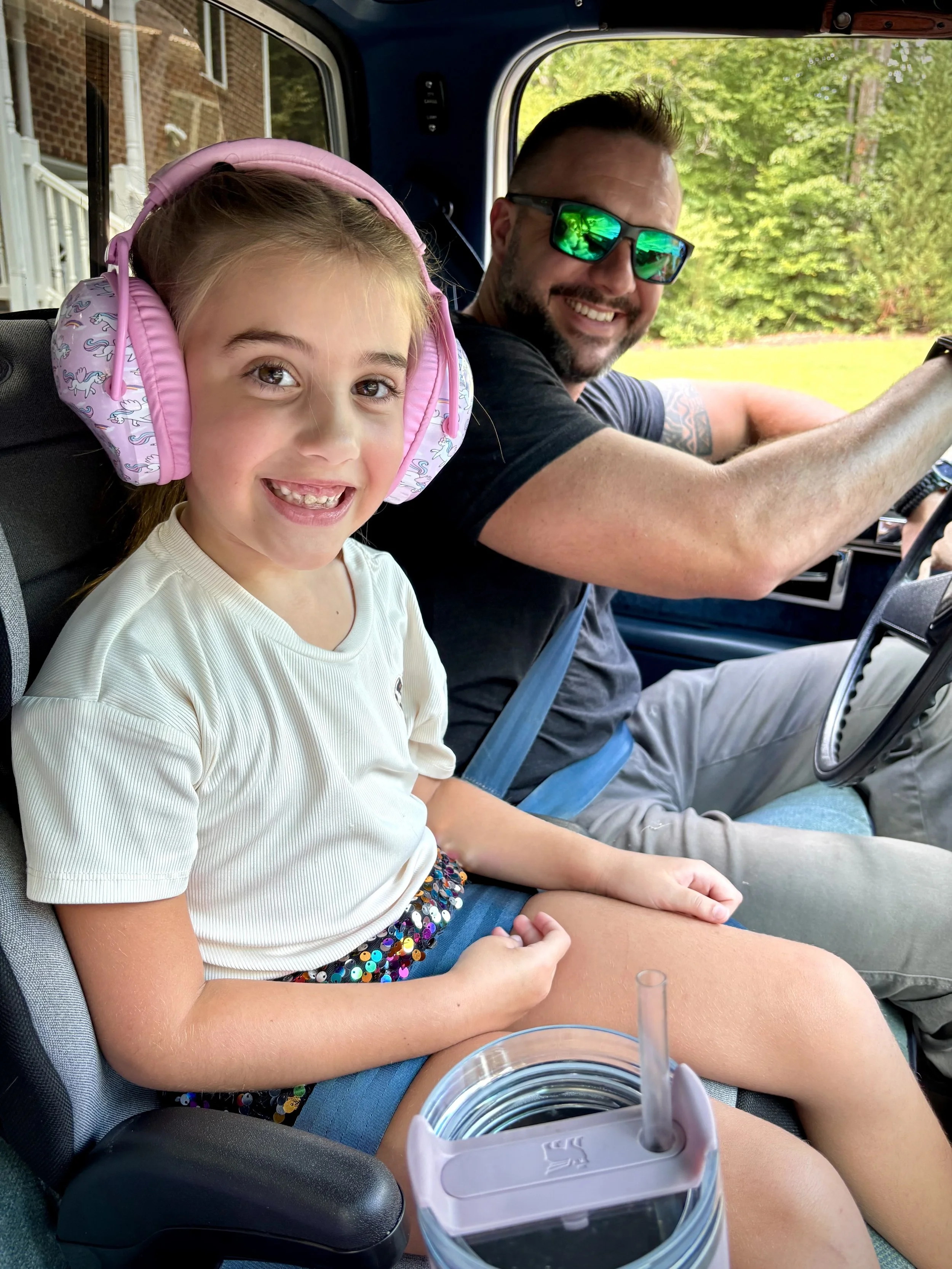 A young girl with brown hair and big brown eyes smiling while wearing pink headphones, sitting in a vehicle with a man, both enjoying a sunny day outdoors with green trees visible through the window.
