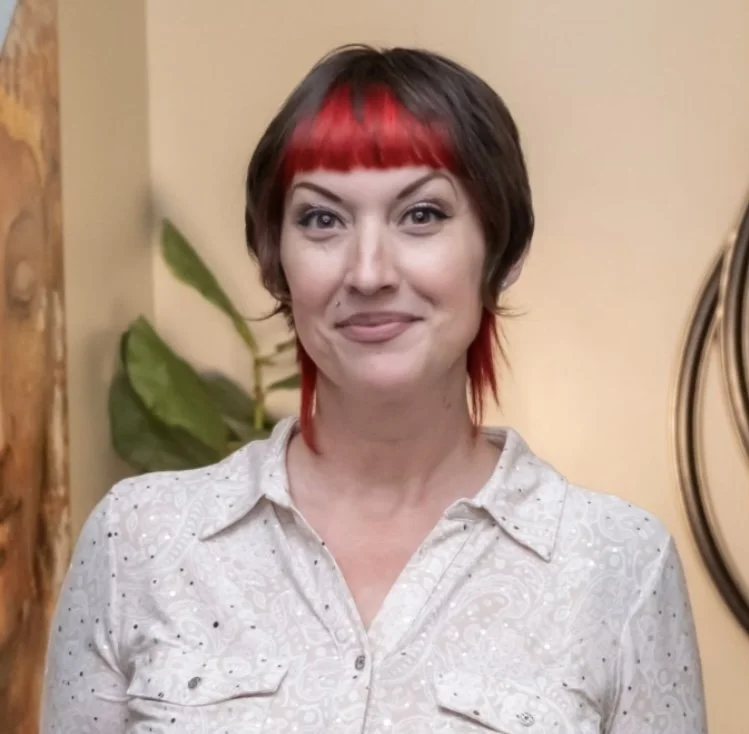 A woman with short, dark hair with red highlights and bangs, wearing a white blouse with embroidery, standing indoors against a beige wall with a houseplant and wooden decor in the background.
