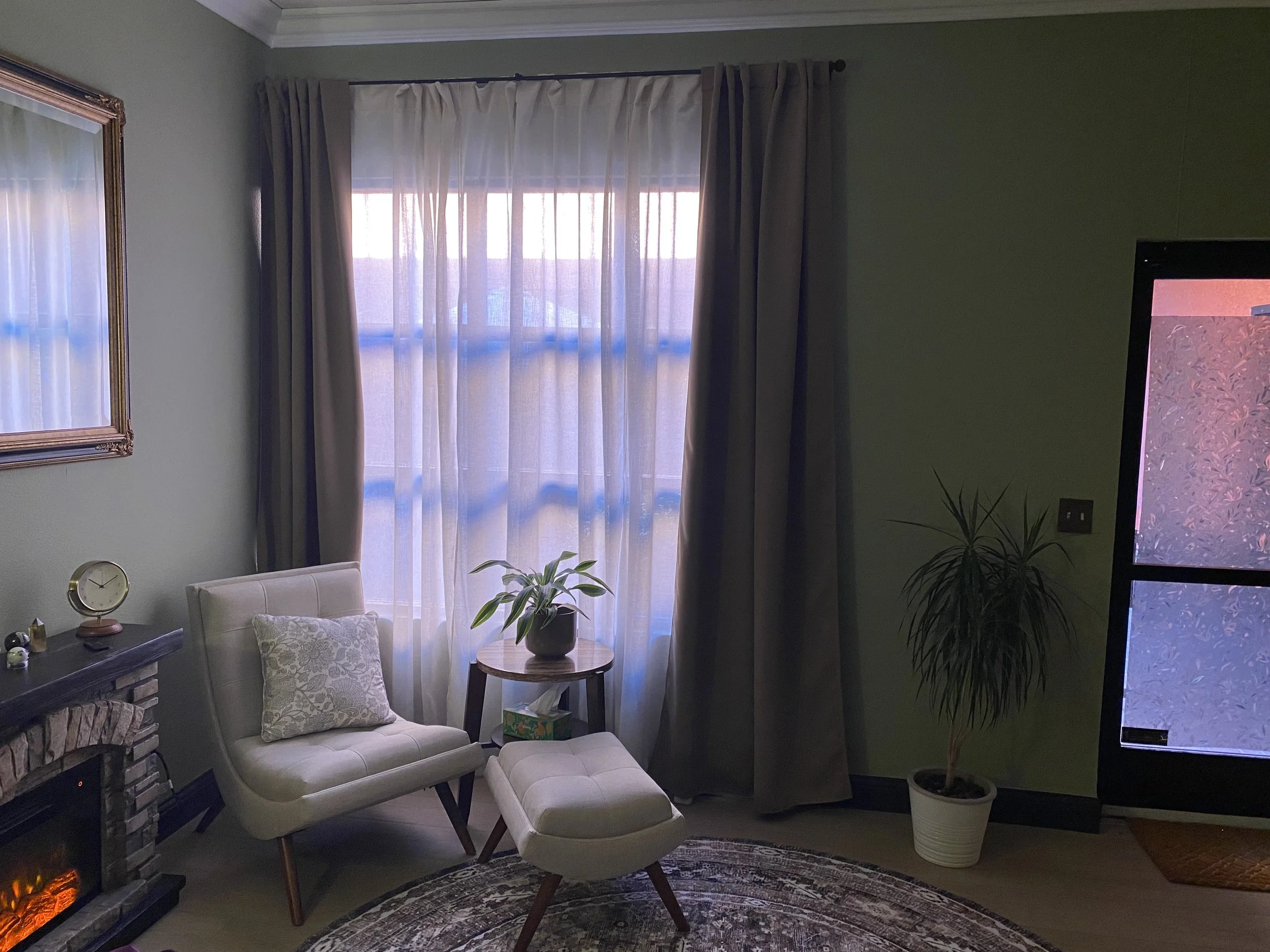 A cozy living room corner with a white armchair, matching ottoman, and a small dresser with a clock on top. There is a window with curtains, a potted plant, and a flat-screen TV on the right side. A decorative rug is on the floor.