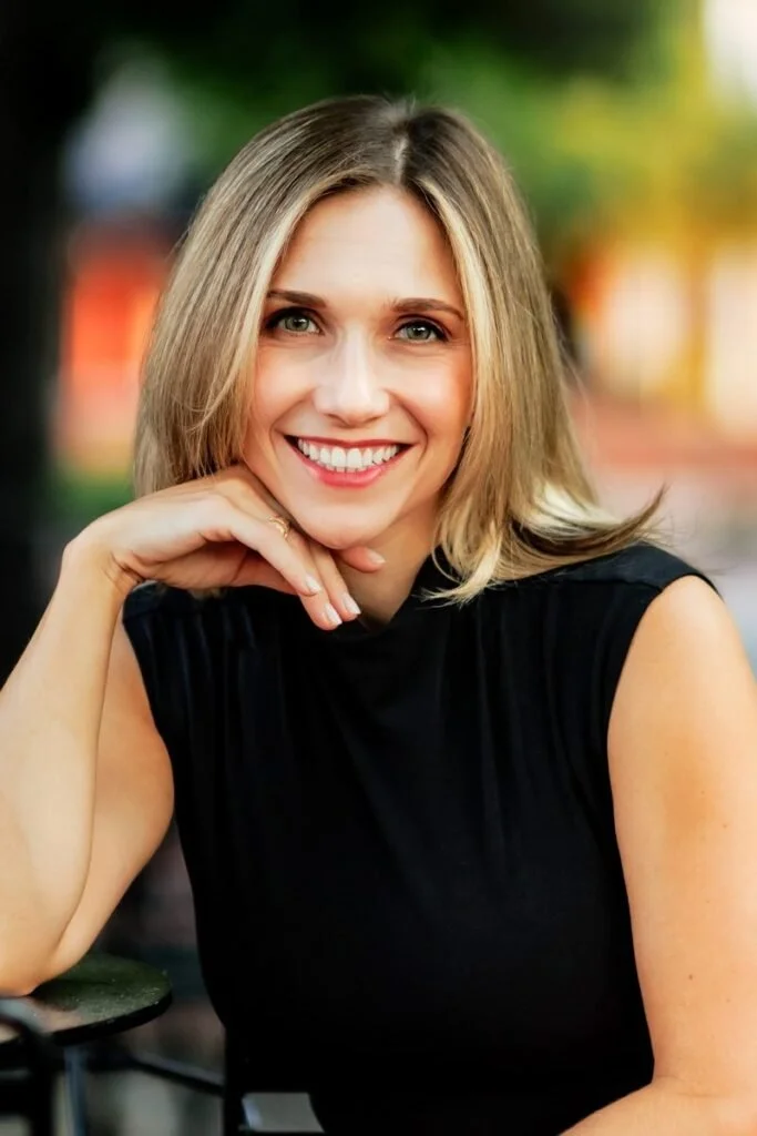 A woman with blonde hair smiling and resting her chin on her hand outdoors during daytime. Elizabeth Ballinger