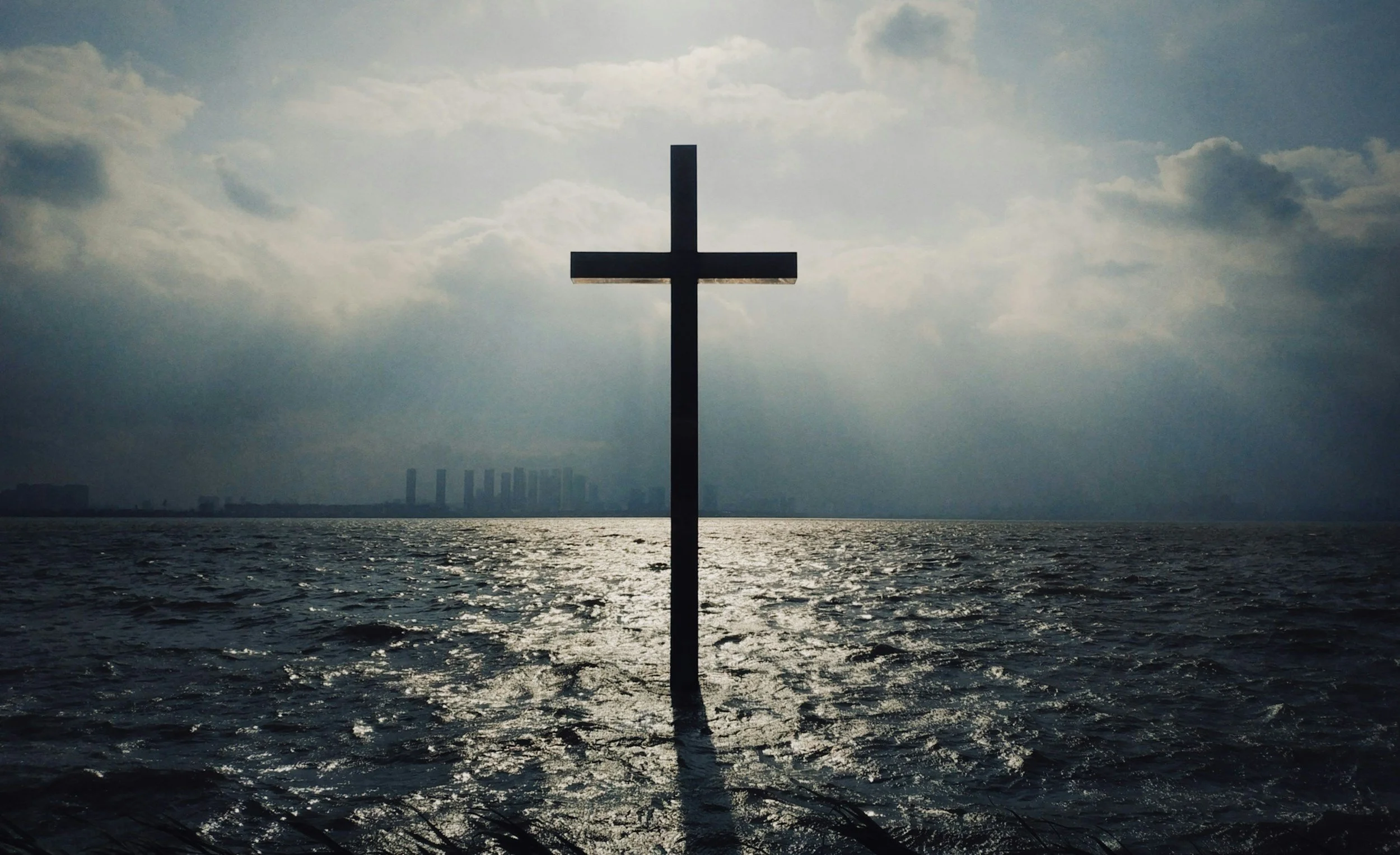 A cross standing in water with a cloudy sky and city skyline in the background.