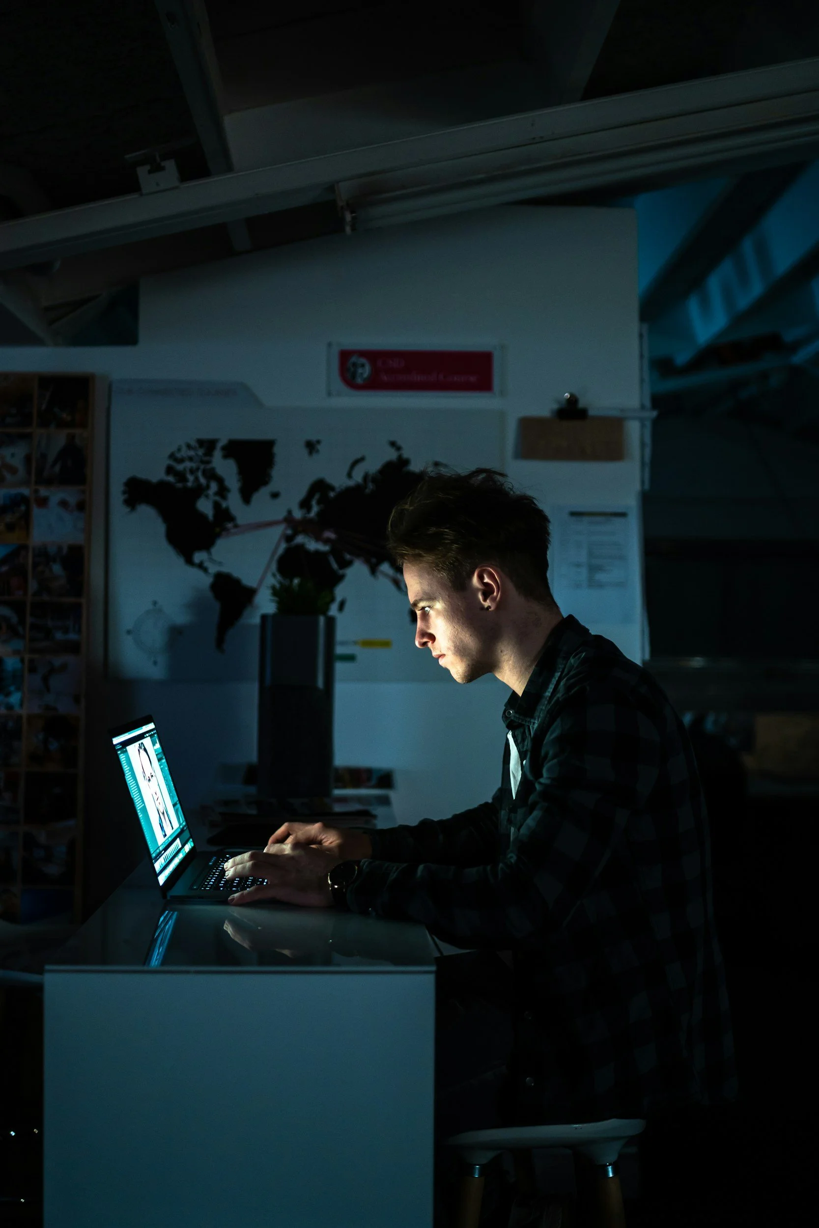 A young man with short dark hair working on a laptop at a desk in a dimly lit room, with a world map on the wall and a sign that reads 'National Geographic' in the background.