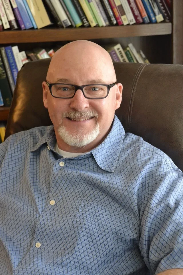 A middle-aged man with a beard and glasses, wearing a light blue checkered shirt, sitting in a leather chair in front of a bookshelf filled with various books. Steve Tryling