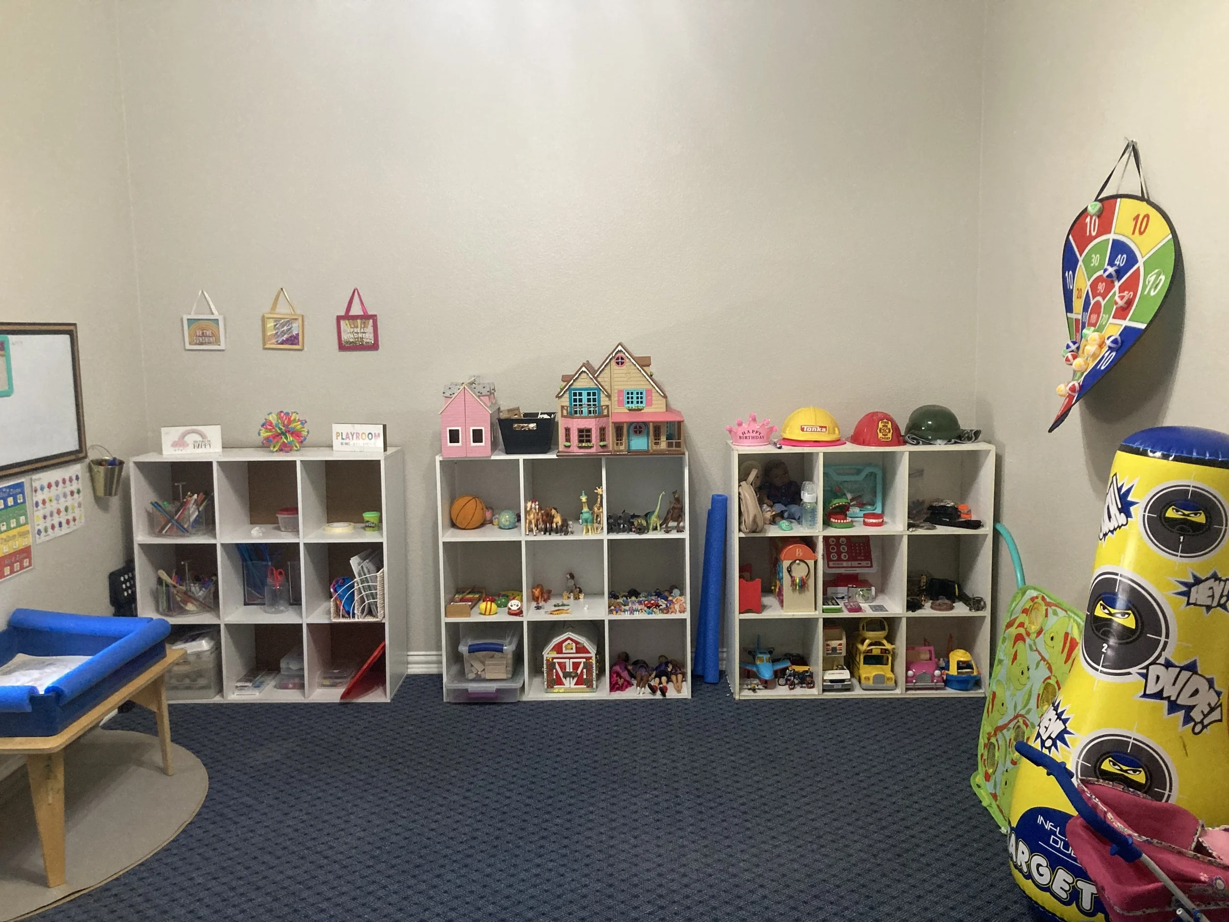 A wall with shelves filled with toys, small houses, and decor items in a playroom or kids' room. There are miniature toy animals, cars, and pictures hanging on the wall. Child Centered play therapy room