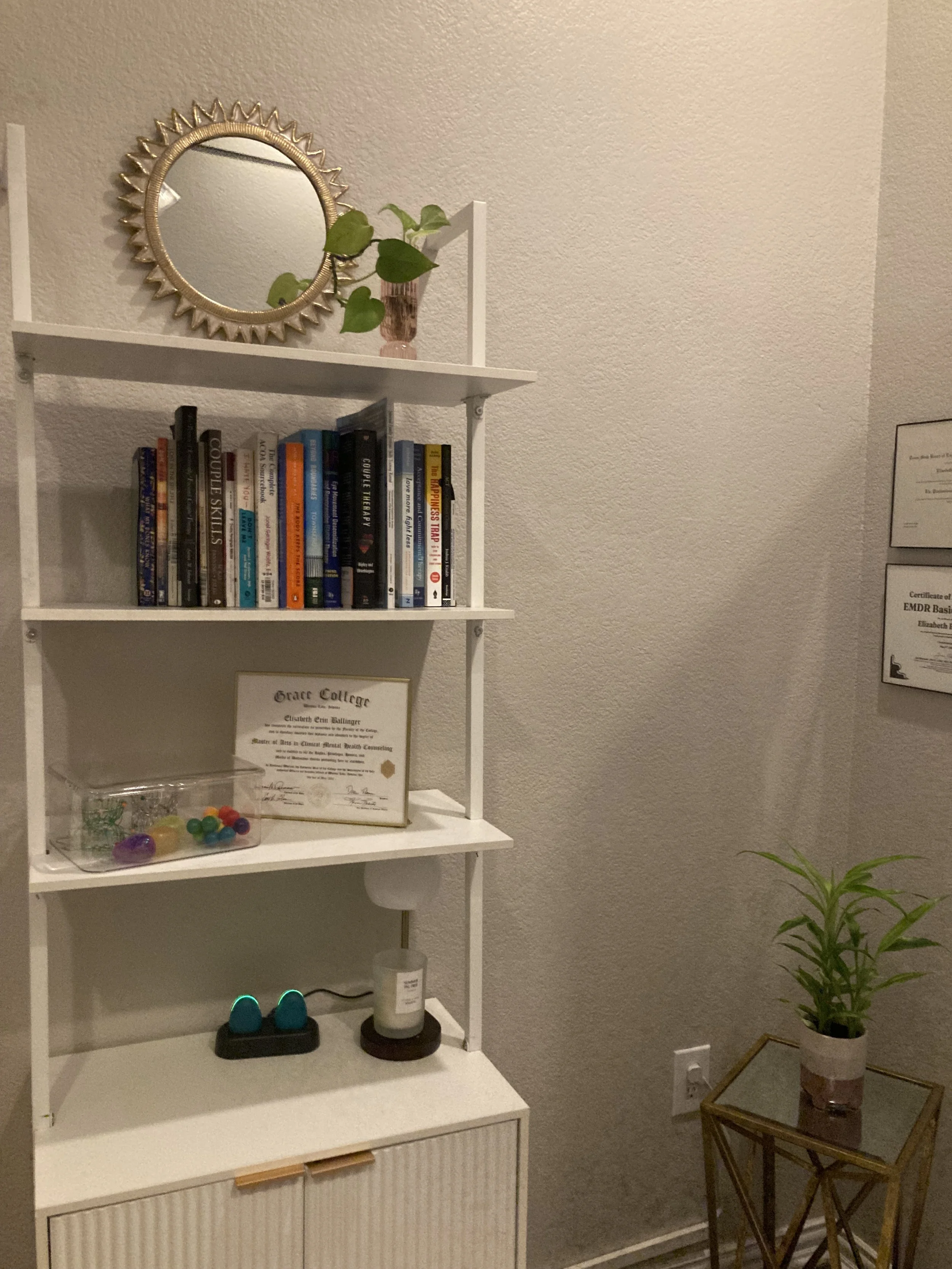 A white bookshelf with three shelves holding books, a glass container with colorful plastic balls, and a framed certificate. On top of the bookshelf is a small potted plant and a circular mirror with a gold sunburst frame. To the right of the bookshelf is a small side table with a tall pink vase and a houseplant. The background features a textured wall and a corner with certificate frames.