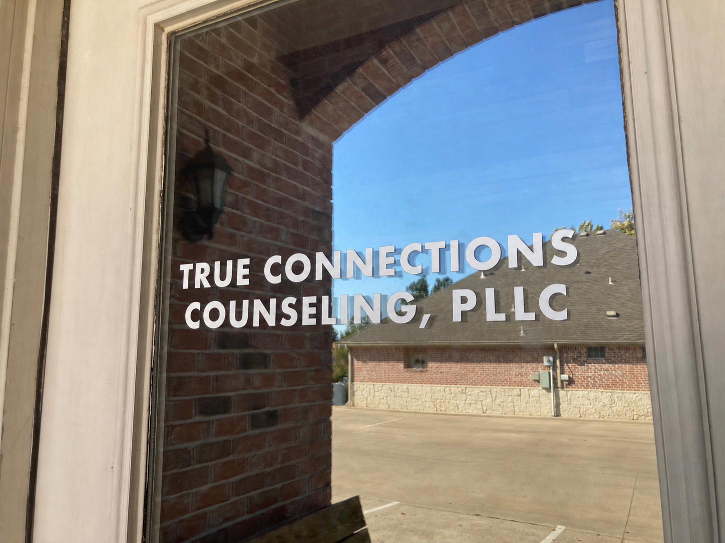 Reflection of a brick building with a gray roof and a parking lot seen in a window with the sign 'True Connections Counseling, PLLC'.