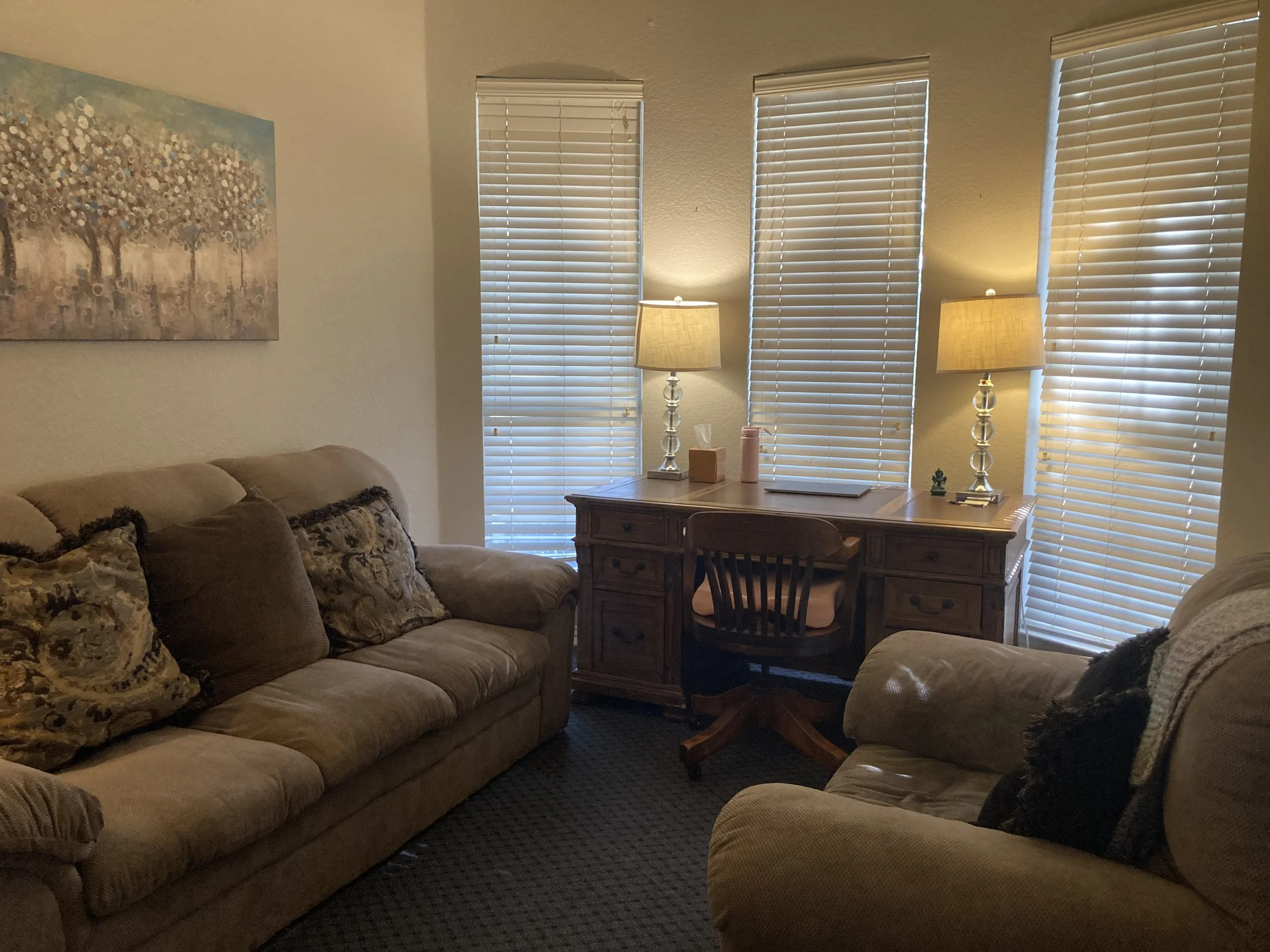 A cozy living room displaying a beige sofa with patterned cushions, a wooden desk with a chair beneath three white window blinds, two table lamps on the desk, and a colorful abstract painting of trees on the wall.