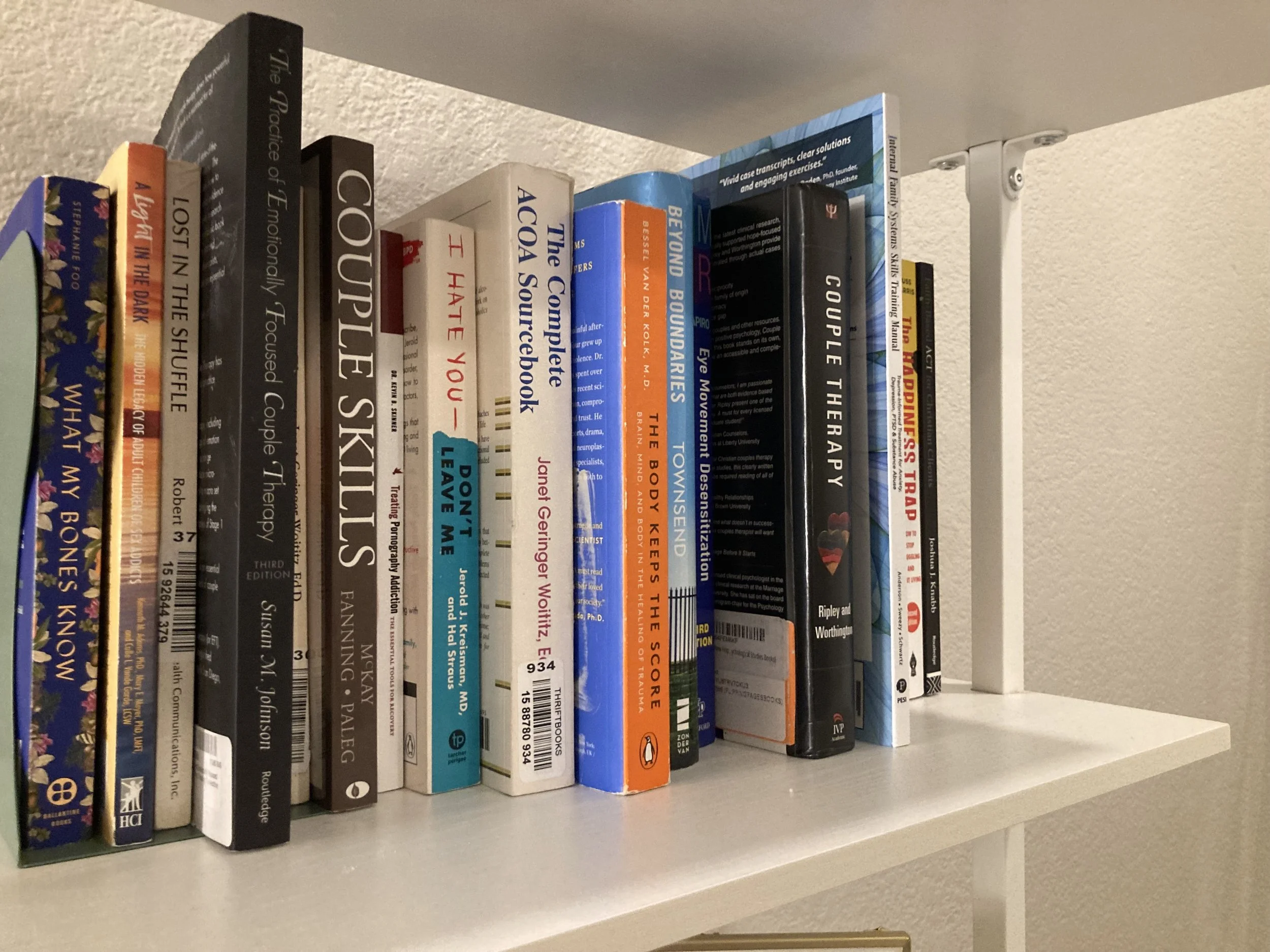 A white bookshelf with a row of various hardcover and paperback books stacked vertically, with some titles including 'The Practice of Emotionally Focused Couple Therapy' and 'Coulple Therapy'.
