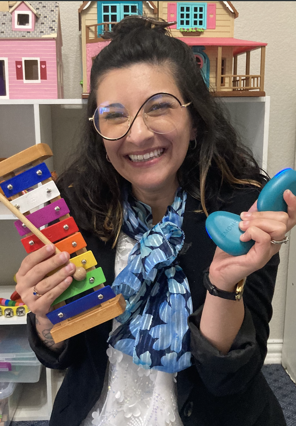 A woman with glasses smiling, holding a colorful xylophone in one hand and a blue stress ball in the other, with a background of dollhouses. Sydney Pimentel-Rushing