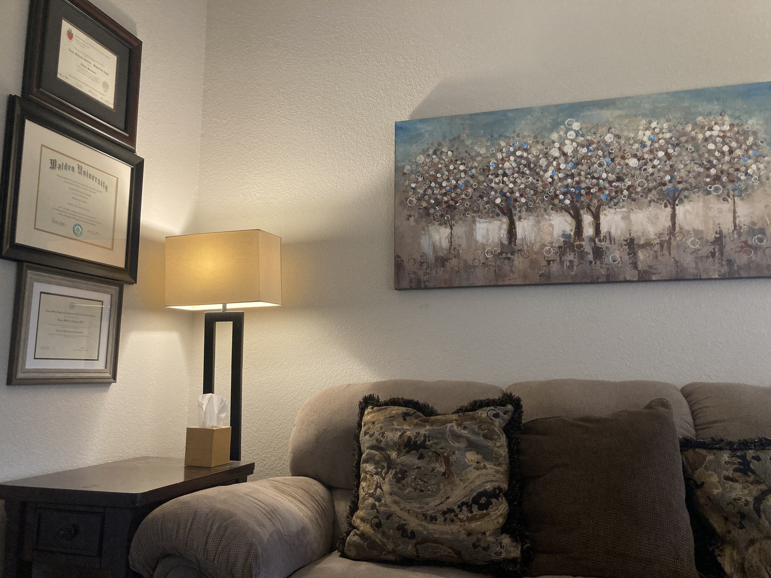 Living room corner with framed diplomas and certificates on the wall, a table lamp on a side table, a beige bookshelf, a large abstract painting of trees, a beige sofa with patterned pillows.