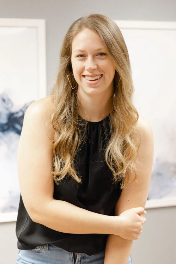 A woman with long, wavy blonde hair smiling, wearing a black sleeveless top and blue jeans, standing indoors with artwork in the background. Jenna Banda
