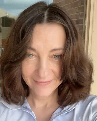 Close-up of a woman with shoulder-length brown hair, blue eyes, smiling, wearing a light blue top, standing outside near a brick wall and window. Laura Cheung