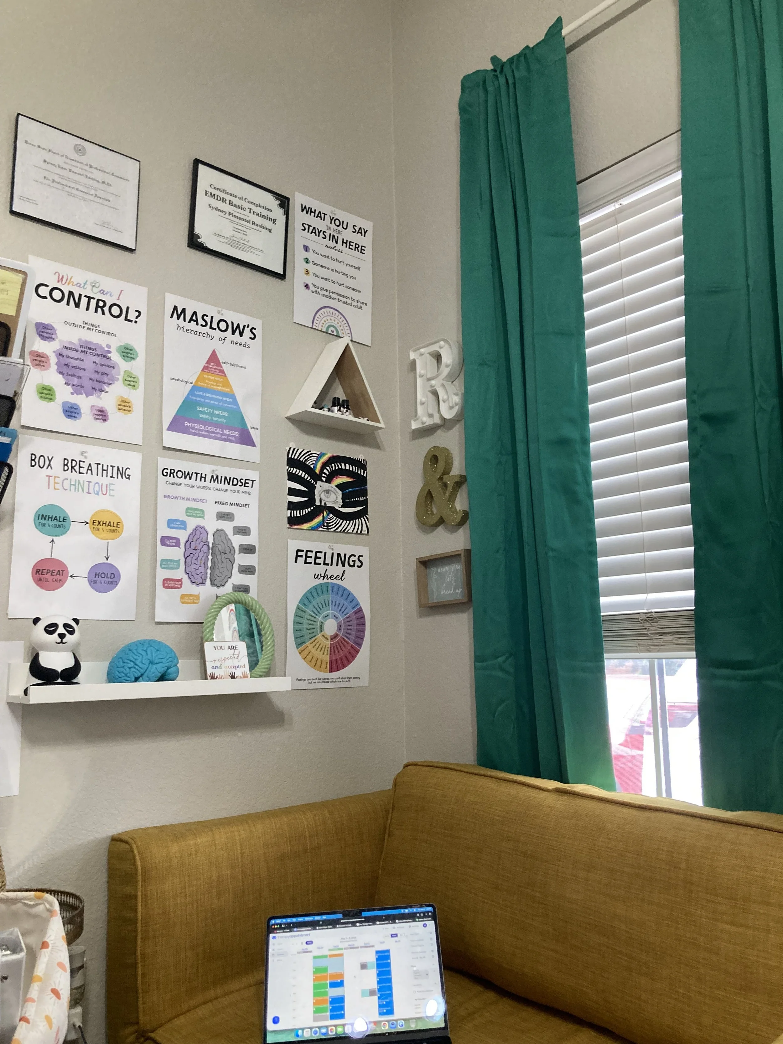 A cozy corner of a room with teal curtains and white window blinds. The wall has various educational and motivational posters, including maslow's hierarchy of needs, feelings wheel, and control chart. A gold ampersand decoration and white shelf with small items are on the wall. A brown upholstered couch and a tablet displaying a colorful calendar or schedule are in the foreground.