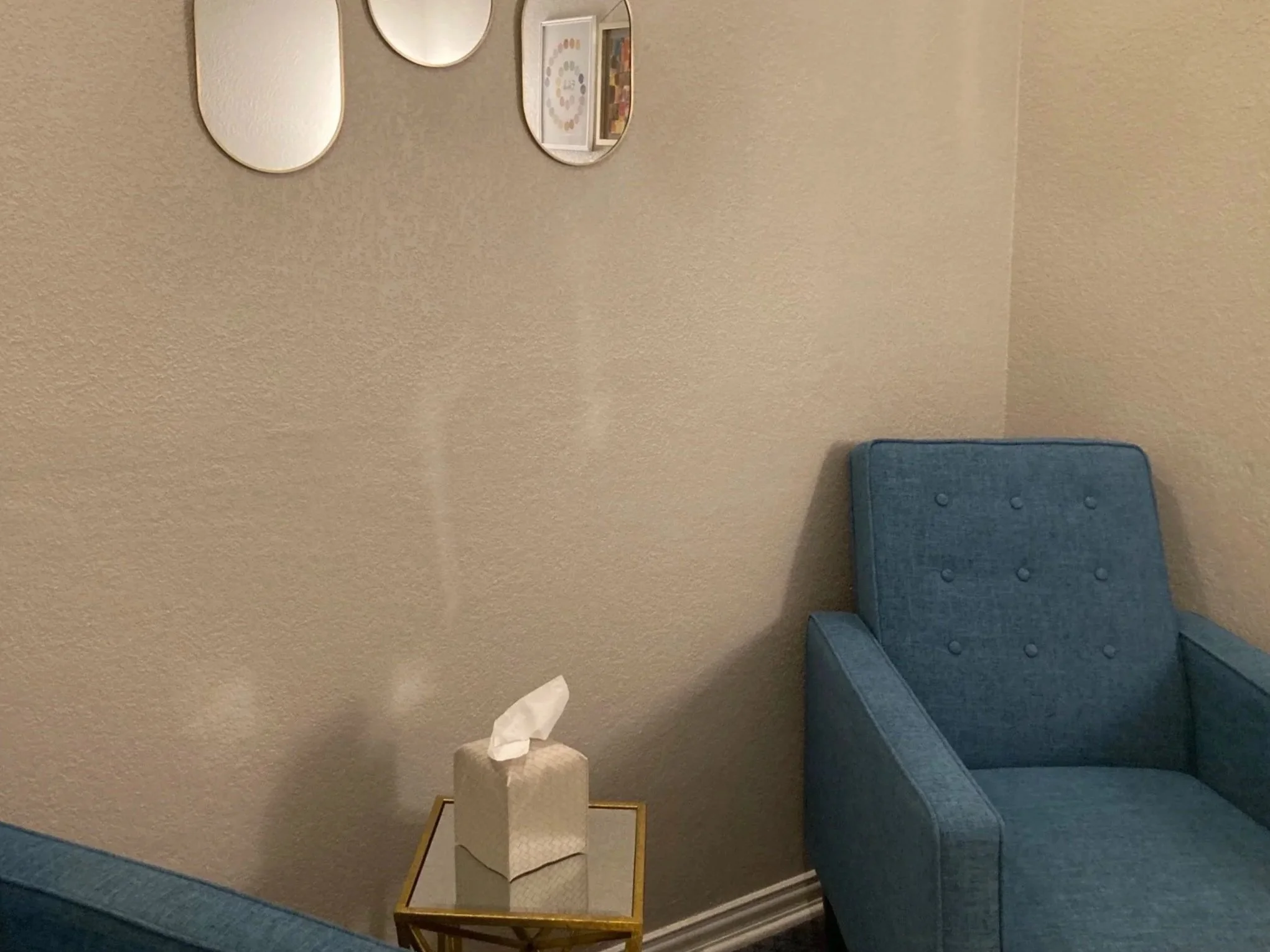 A small side table with a crumpled tissue on top, next to a blue upholstered chair in a room with beige textured walls, a mirror, framed pictures, and a gold-framed tray