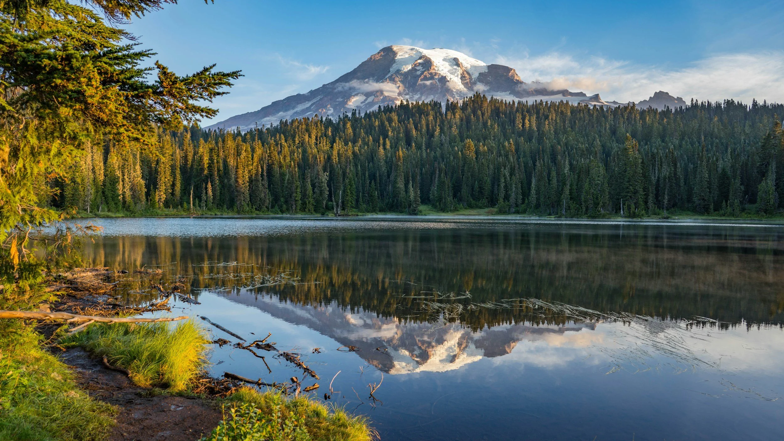 A mountain with snow on top, surrounded by a forest, reflected in a calm lake with green grass and trees along the shoreline.