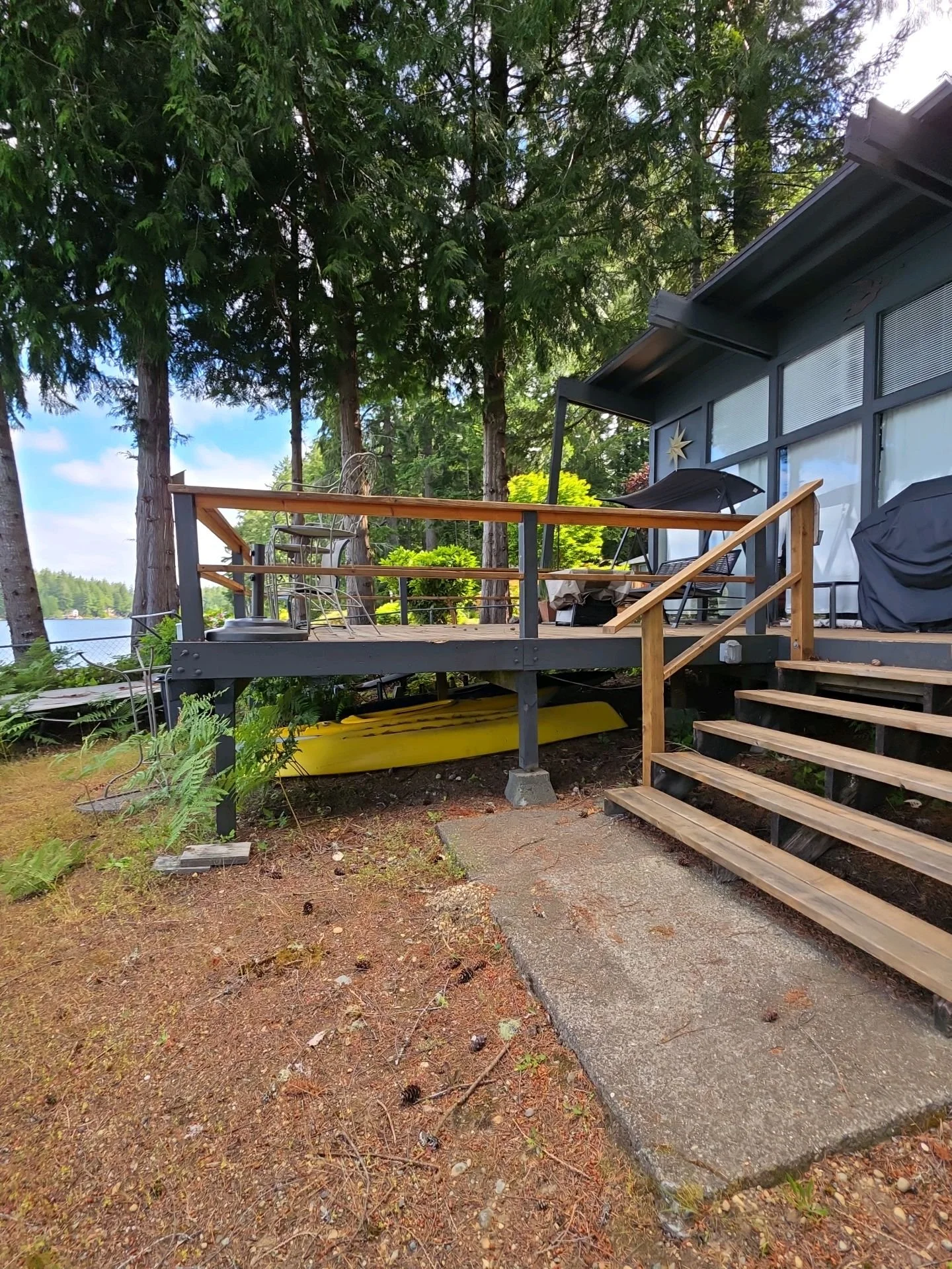 A wooden deck with outdoor furniture and a grill, surrounded by tall trees near a lake, with a partly cloudy sky overhead.