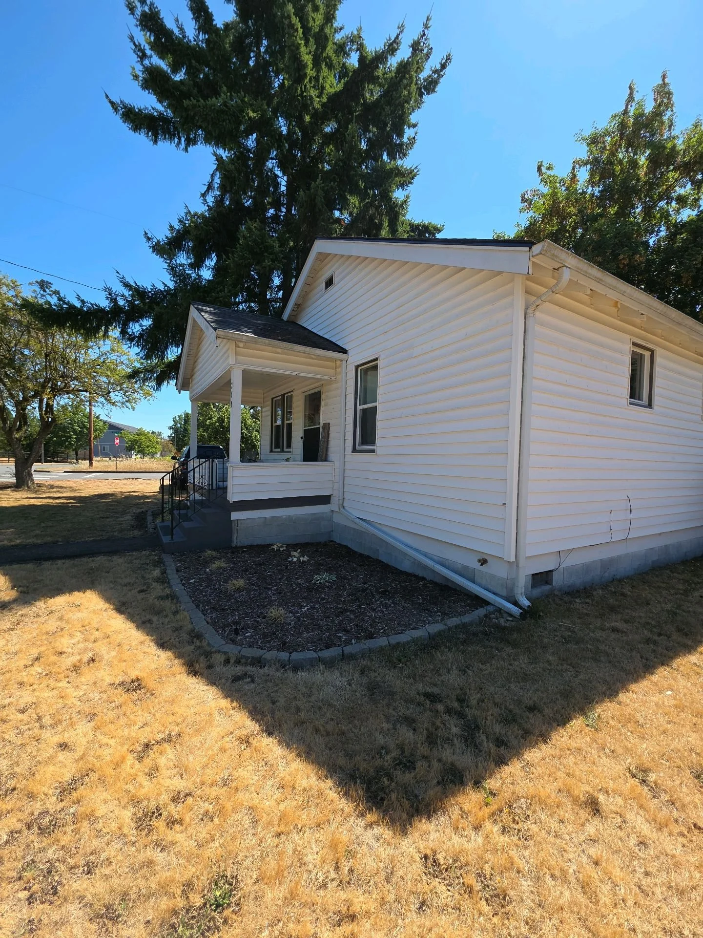 White house with small front porch, black railing, and steps, surrounded by yellowish dry grass and trees, under a clear blue sky.