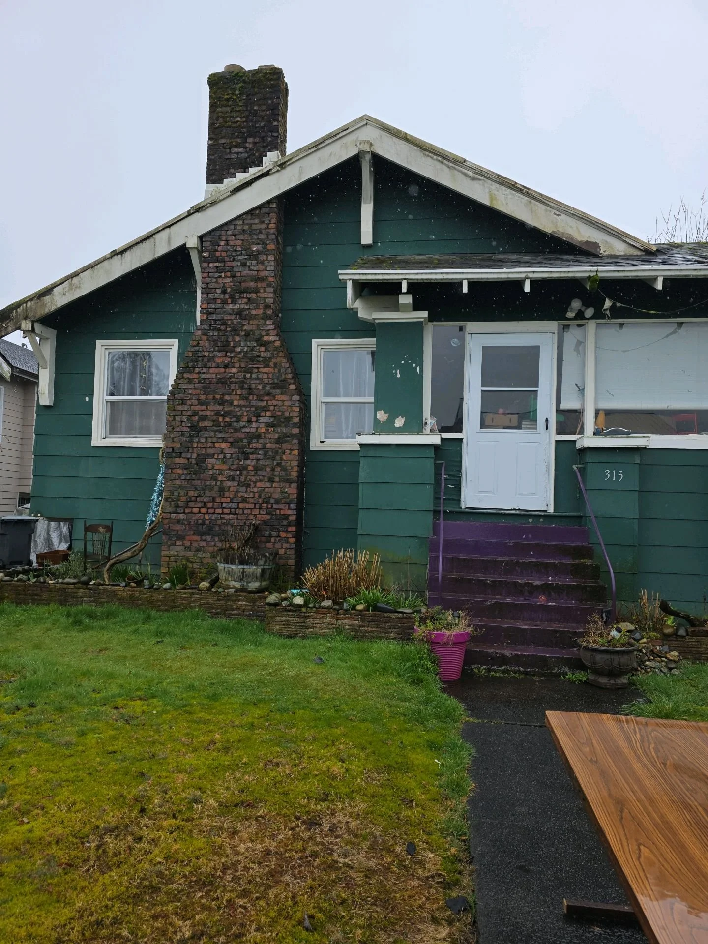 A house with green siding, a brick chimney, and a white front door. The house has a small front porch with purple steps, a purple flower pot, and various plants in garden beds. The lawn is green and well-maintained. The sky is cloudy.