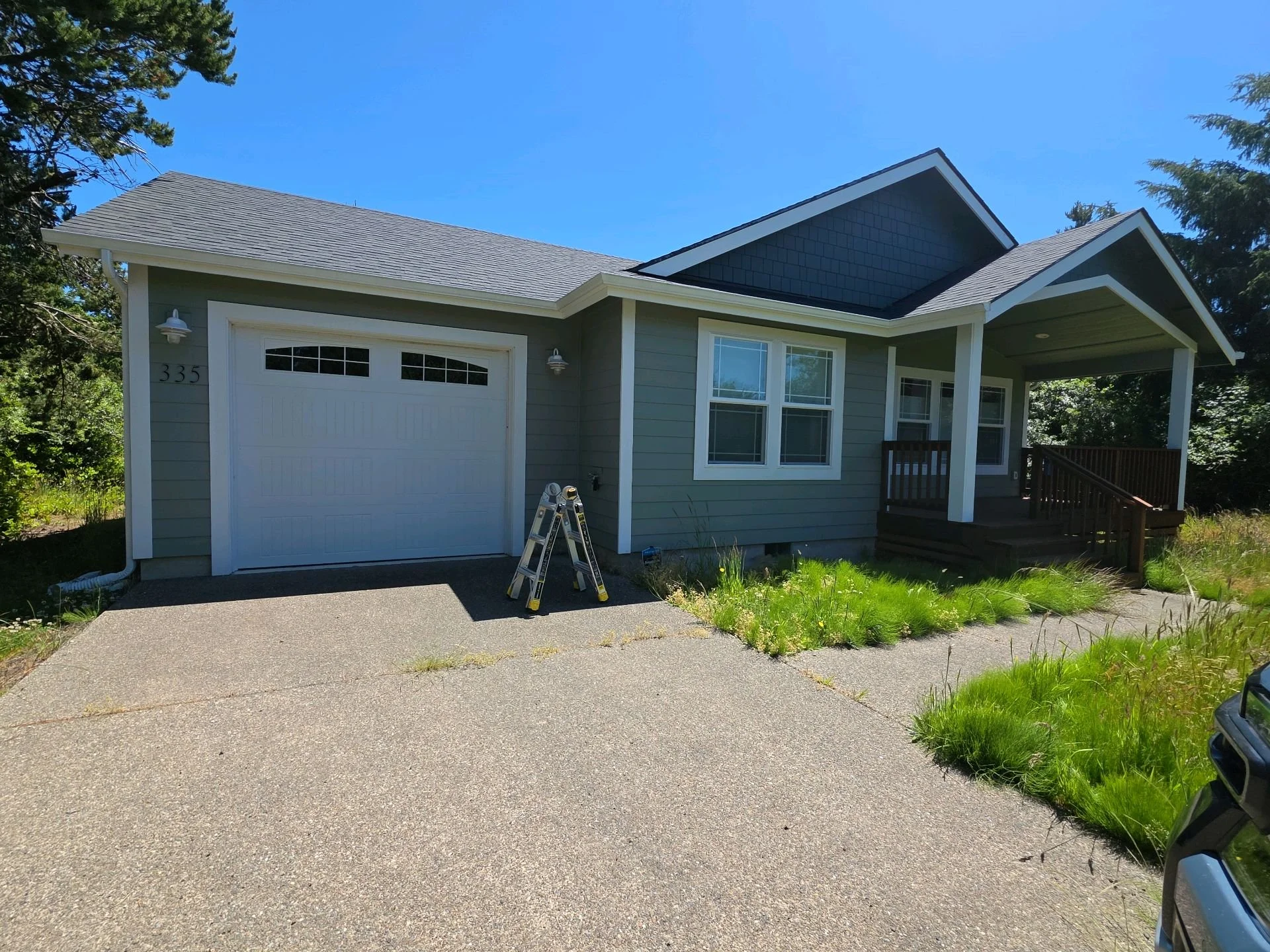 A house with gray siding, a white garage door, and a small porch with a railing. There are trees on both sides of the house and a clear blue sky above.