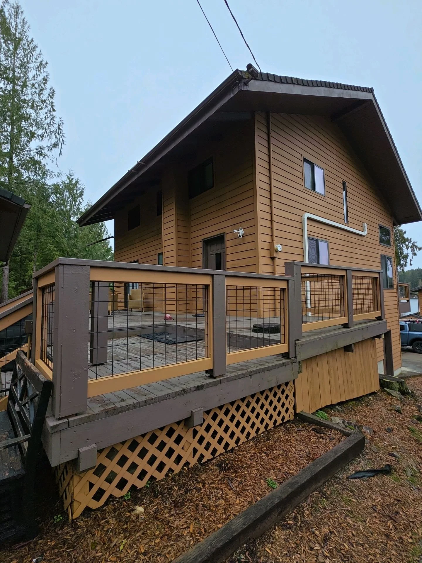 A two-story wooden house with a deck and railing in a natural setting with trees. There is a security camera and a white pipe on the exterior wall.