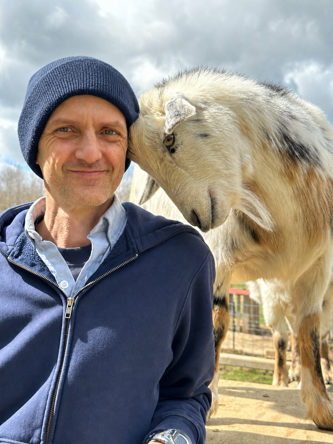 A man in a blue jacket and beanie hat smiling shown close-up with a white and black goat nuzzling his face outdoors under cloudy sky.