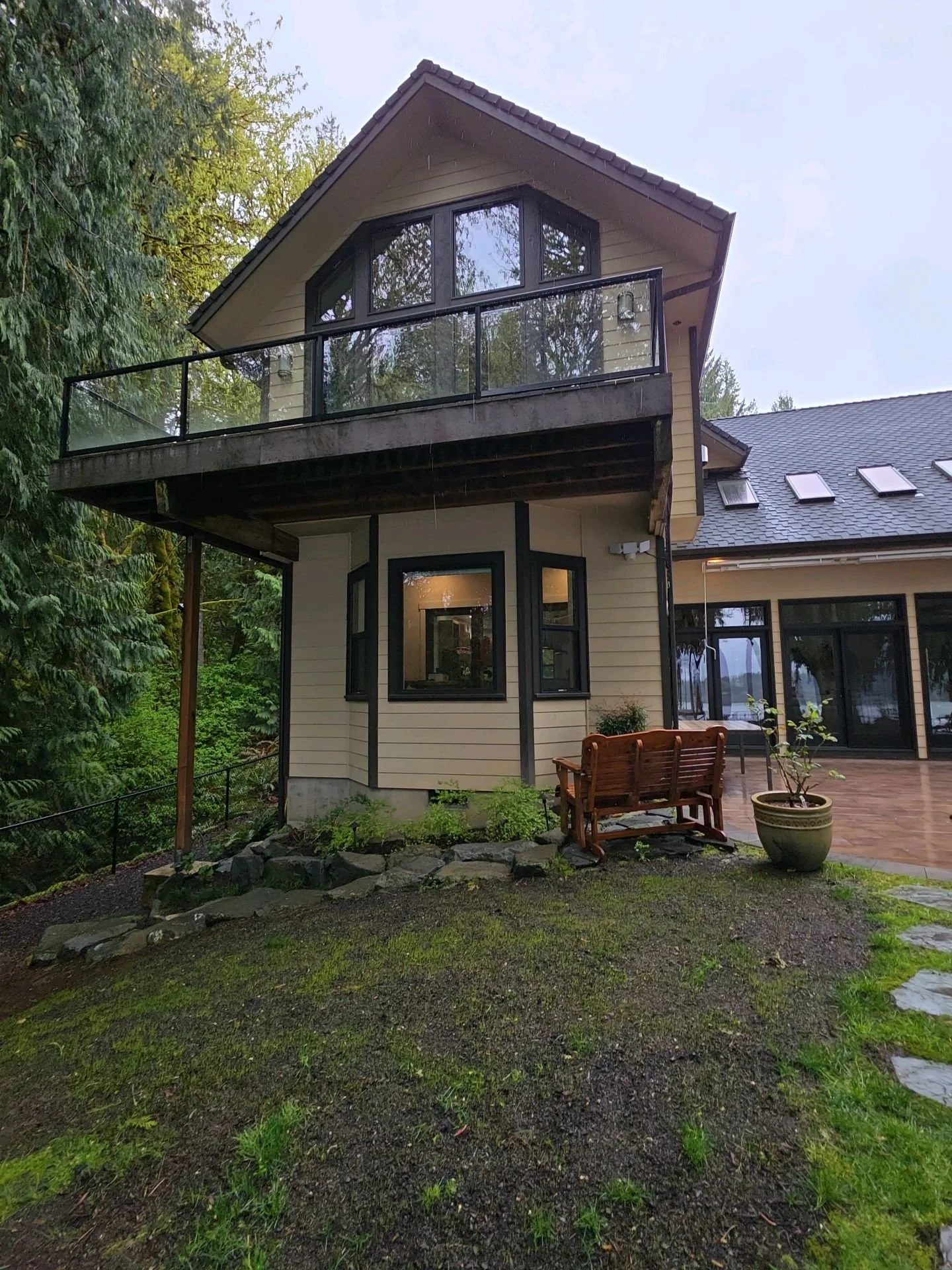 A modern two-story house with large windows and a balcony, surrounded by lush green trees and a garden with a bench and potted plants, during a rainy day.