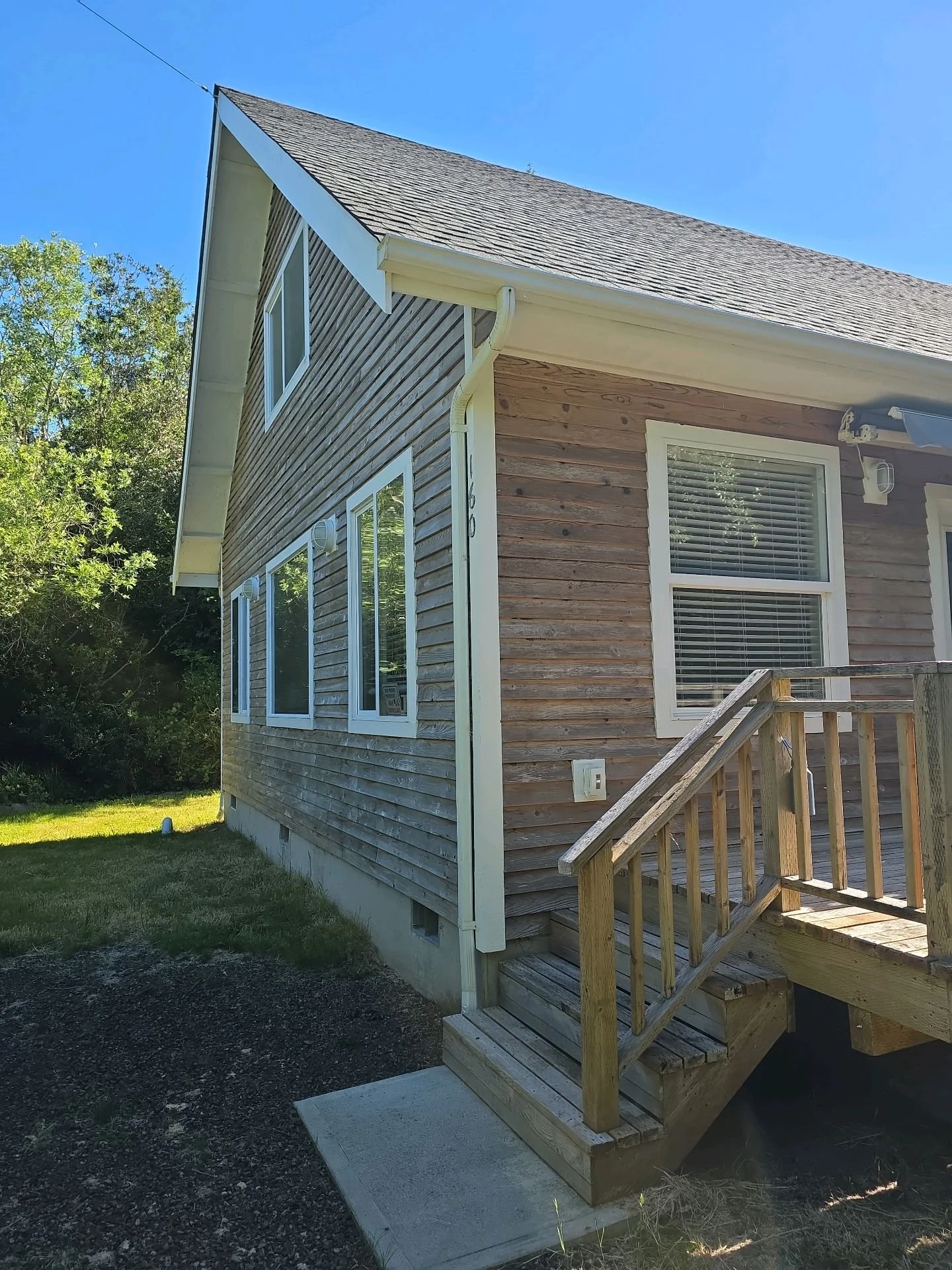 Exterior of a two-story house with wooden siding, white trim, a small staircase, windows, and a pitched roof under a clear blue sky.