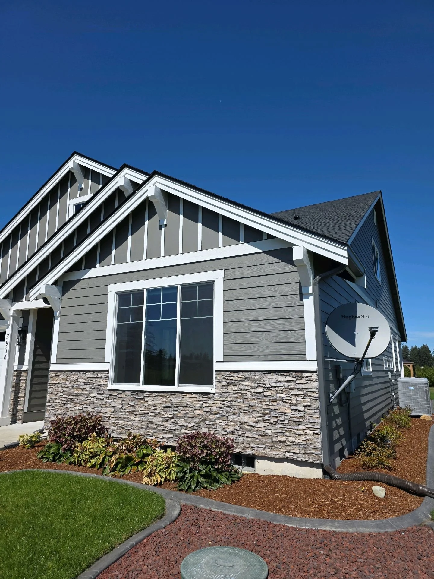 Front corner view of a modern two-story house with gray vinyl siding, stone accents at the base, a large window, and a satellite dish attached to the side. The well-maintained lawn and landscaped garden with plants are visible in the foreground, under a clear blue sky.