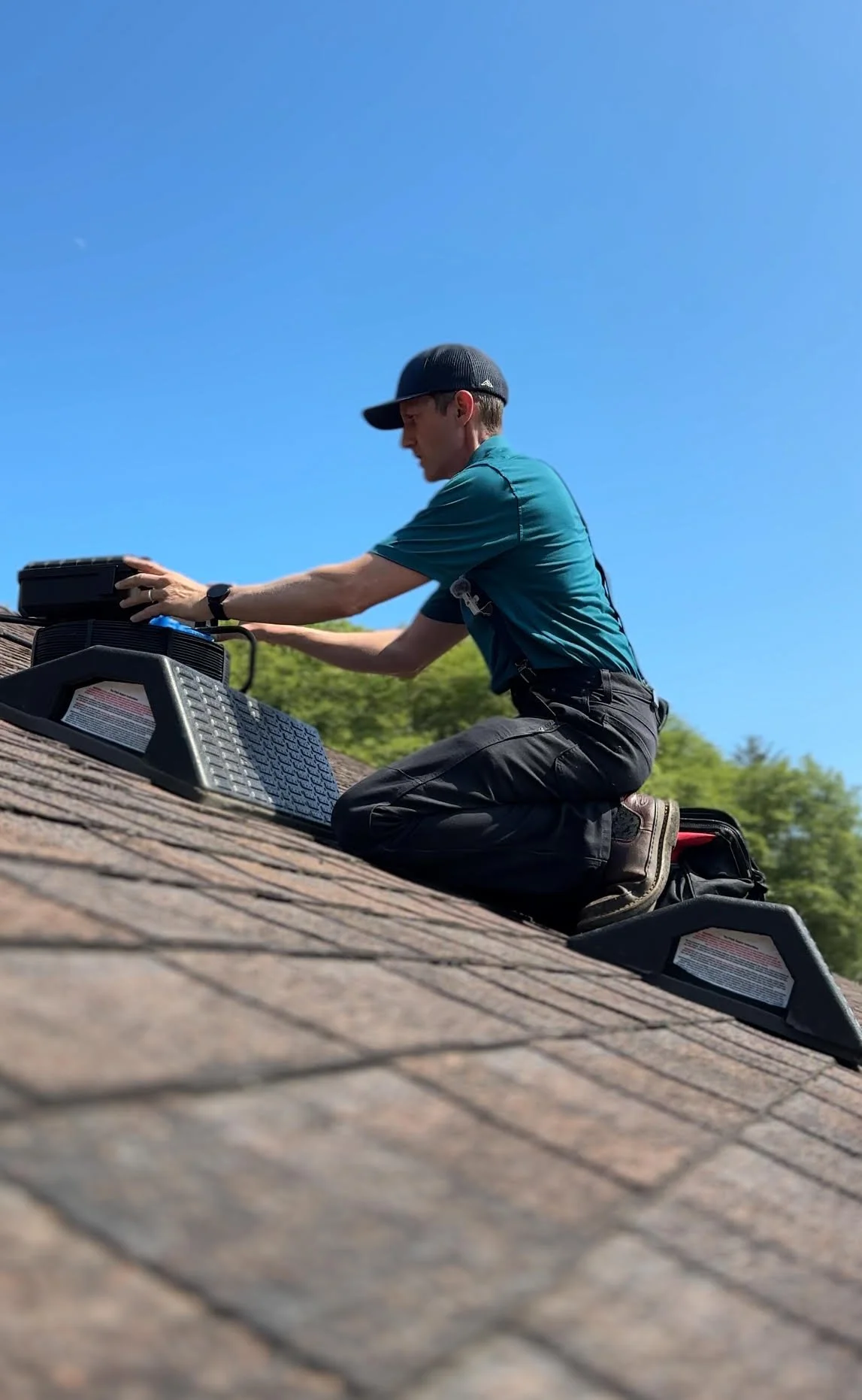A man kneeling on a roof installing or servicing a solar panel against a clear blue sky, with green trees in the background.