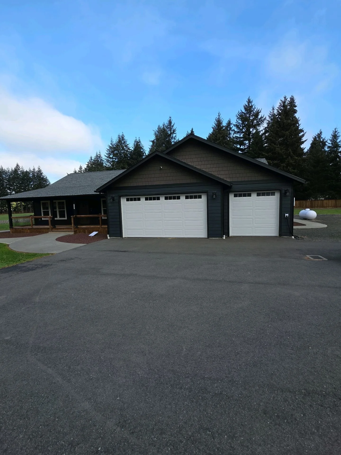 A house with a two-car garage and a porch in a residential neighborhood surrounded by trees. The house has dark siding and white garage doors. The driveway and sidewalk are visible.