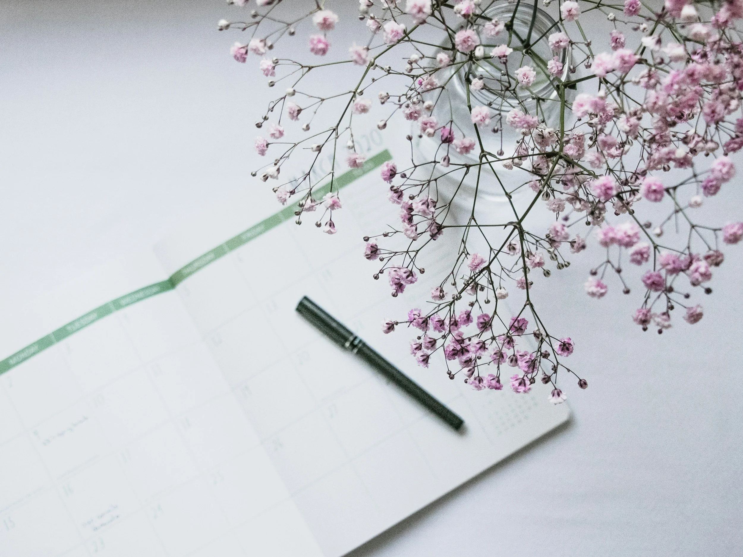 Pink baby's breath flowers in a glass vase placed on an open notebook with a black pen on a white surface.