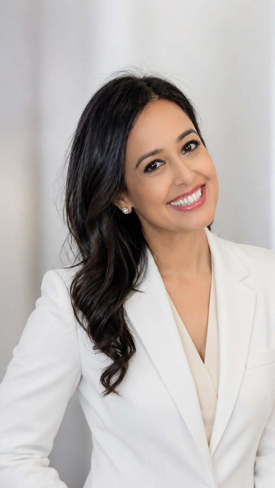 Professional headshot of a woman with long dark wavy hair, smiling, wearing a white blazer and earrings, against a neutral background.