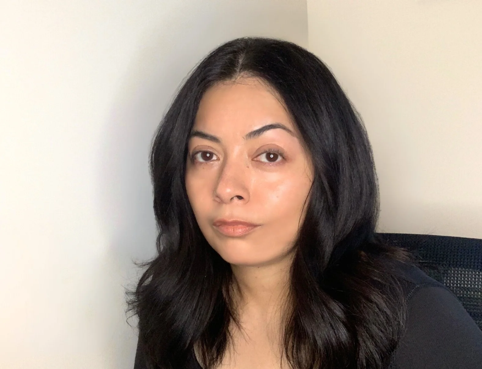 A woman with long black hair, light skin, and wearing a black top, sitting against a plain light-colored wall.