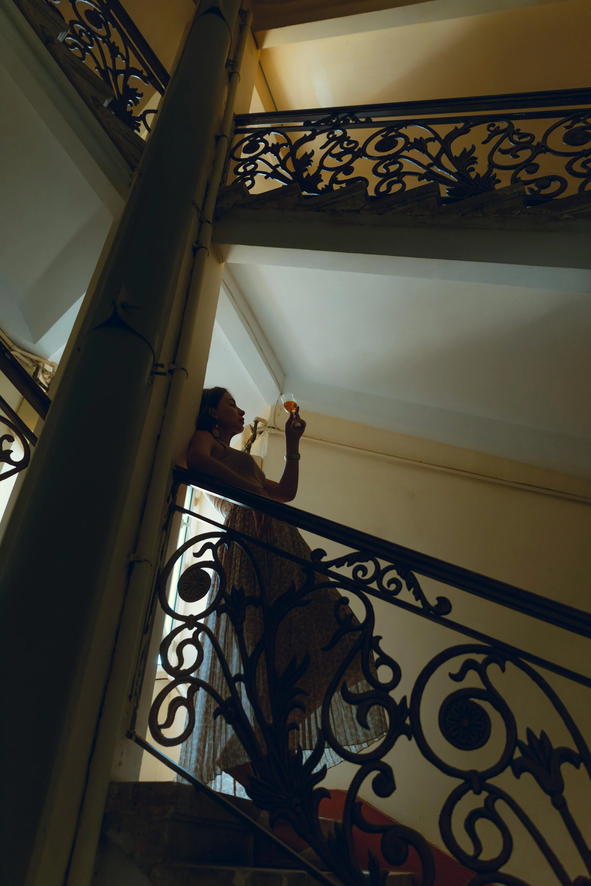 A woman standing on a staircase, holding a glass, seen from below through ornate metal railings.