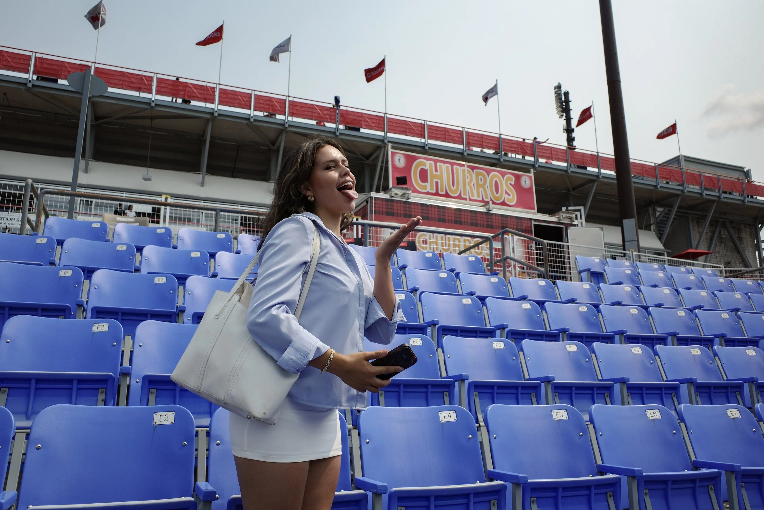 Young woman with brown hair, wearing a white blazer and white skirt, standing in a stadium with bright blue seats, sticking out her tongue and holding a phone.