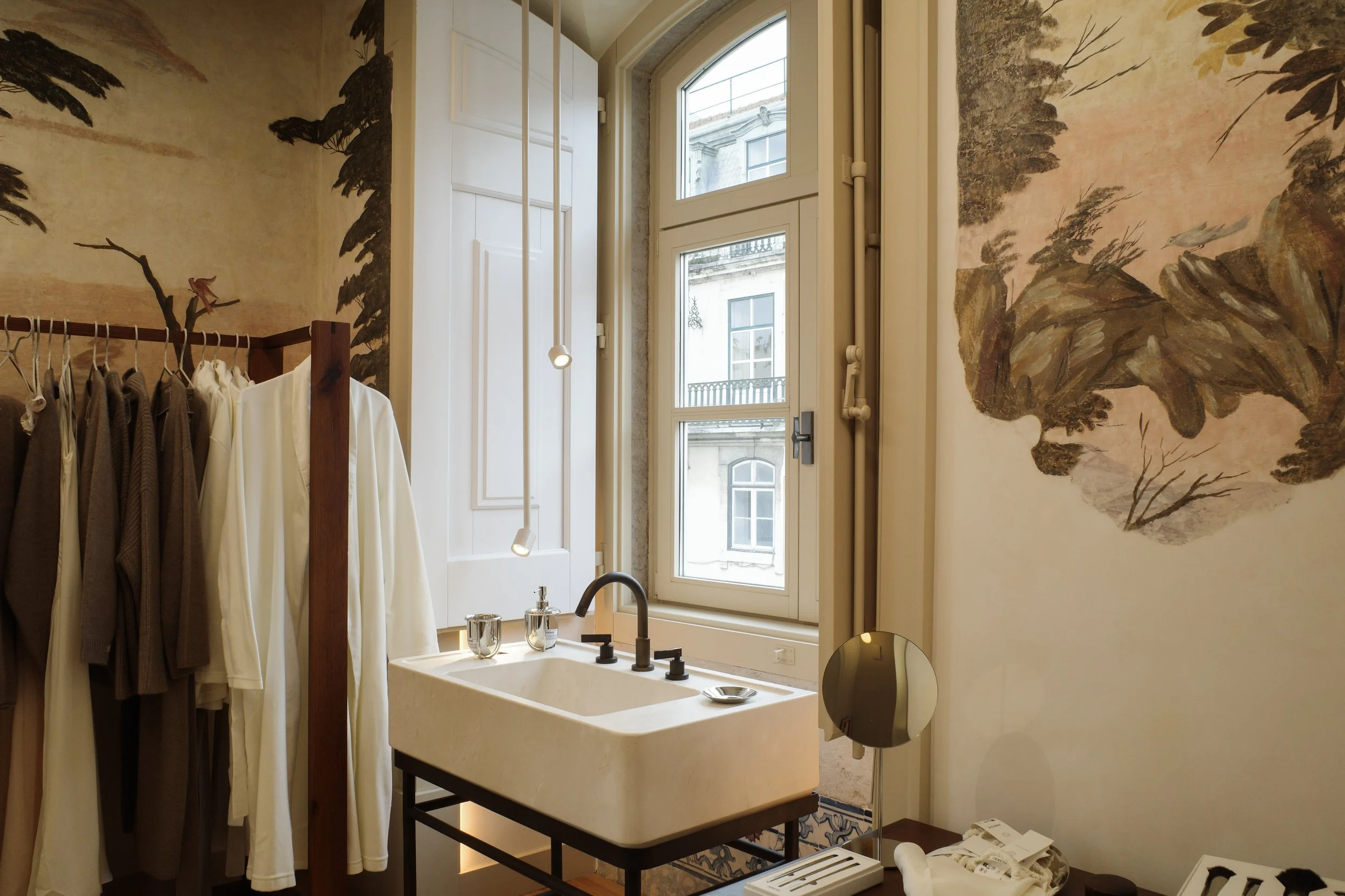 Interior of a room with a large window, white wooden panels, a mural of trees on the wall, and a clothing rack with beige and white garments. A beige sink with a black faucet and some toiletries is visible in the foreground.
