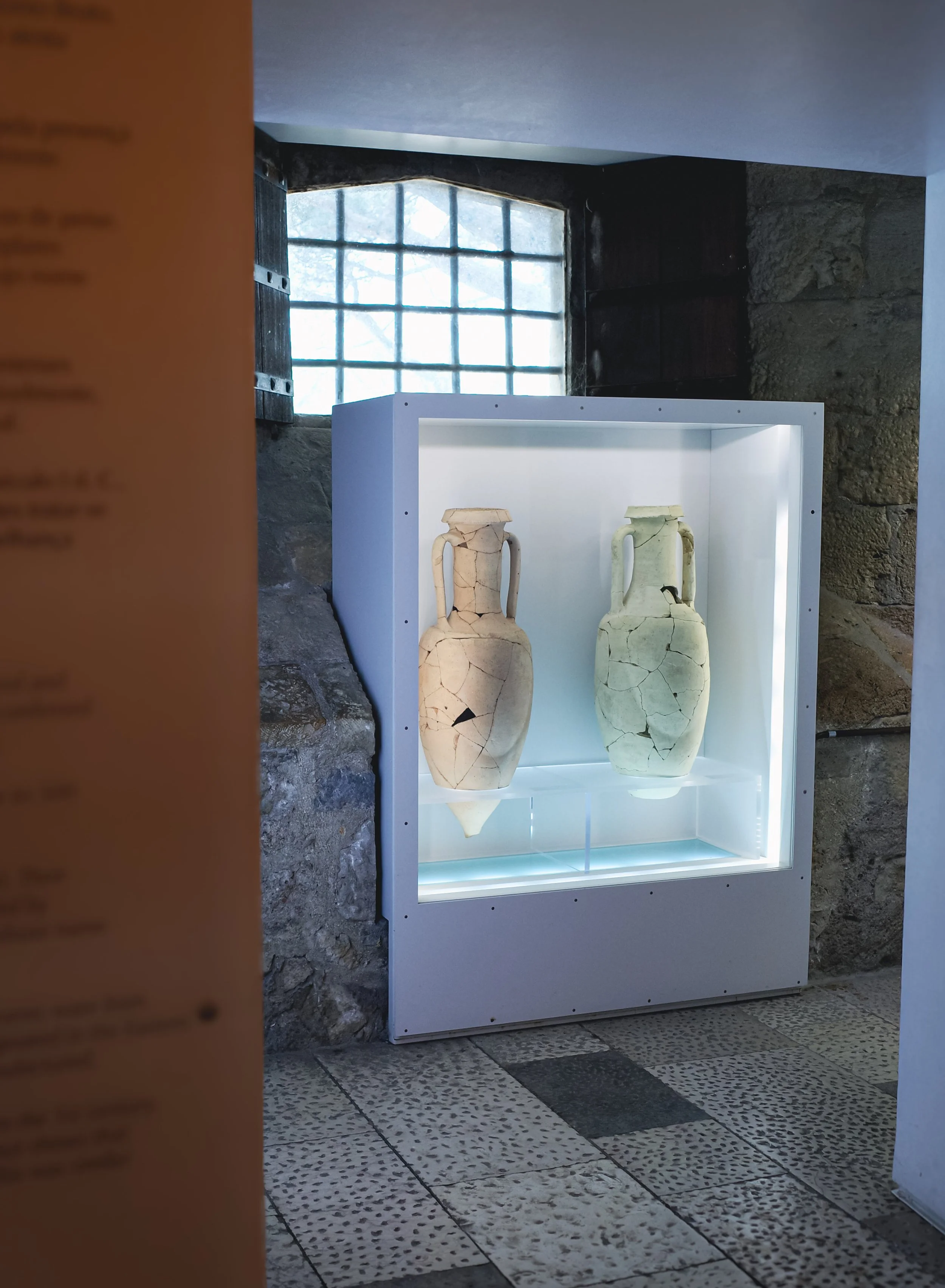 Two ancient pottery vessels with visible cracks, displayed in a modern glass case with white interior lighting, in a stone-walled museum or exhibit space near a window with metal bars.