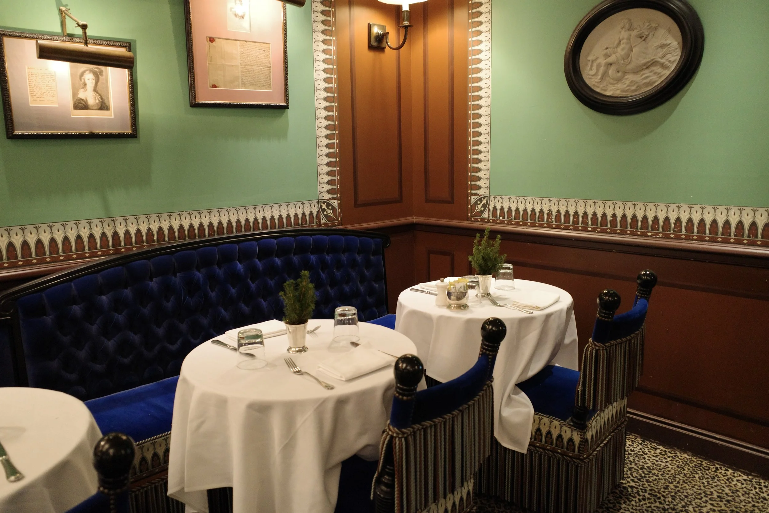 A cozy, decorated restaurant interior with green and pink walls, framed art, a carved wall relief, and tables set with white tablecloths, glassware, silverware, small potted plants, and dark wood chairs with painted fabric seats.