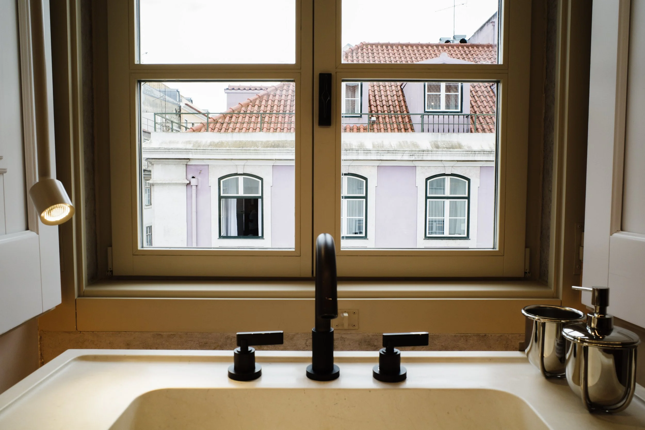 View through a kitchen window showing rooftops with red tiles and a pink building with arched windows inside.
