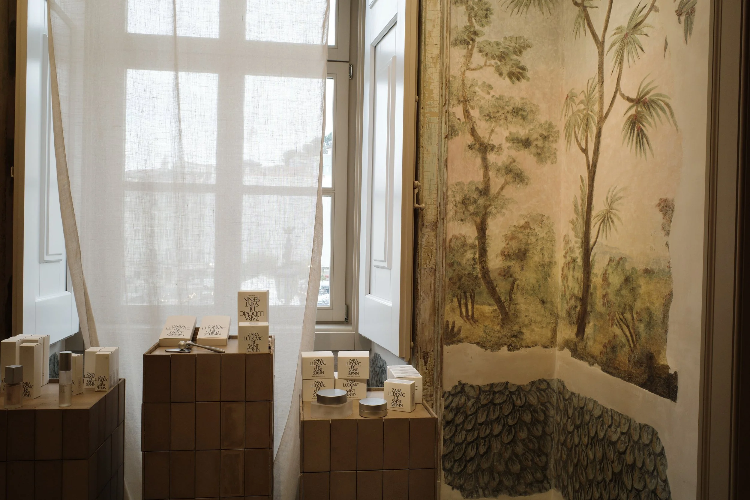 Display of Zara LUDOVIC products with a window and curtain in the background and a mural with trees and plants on the right side.