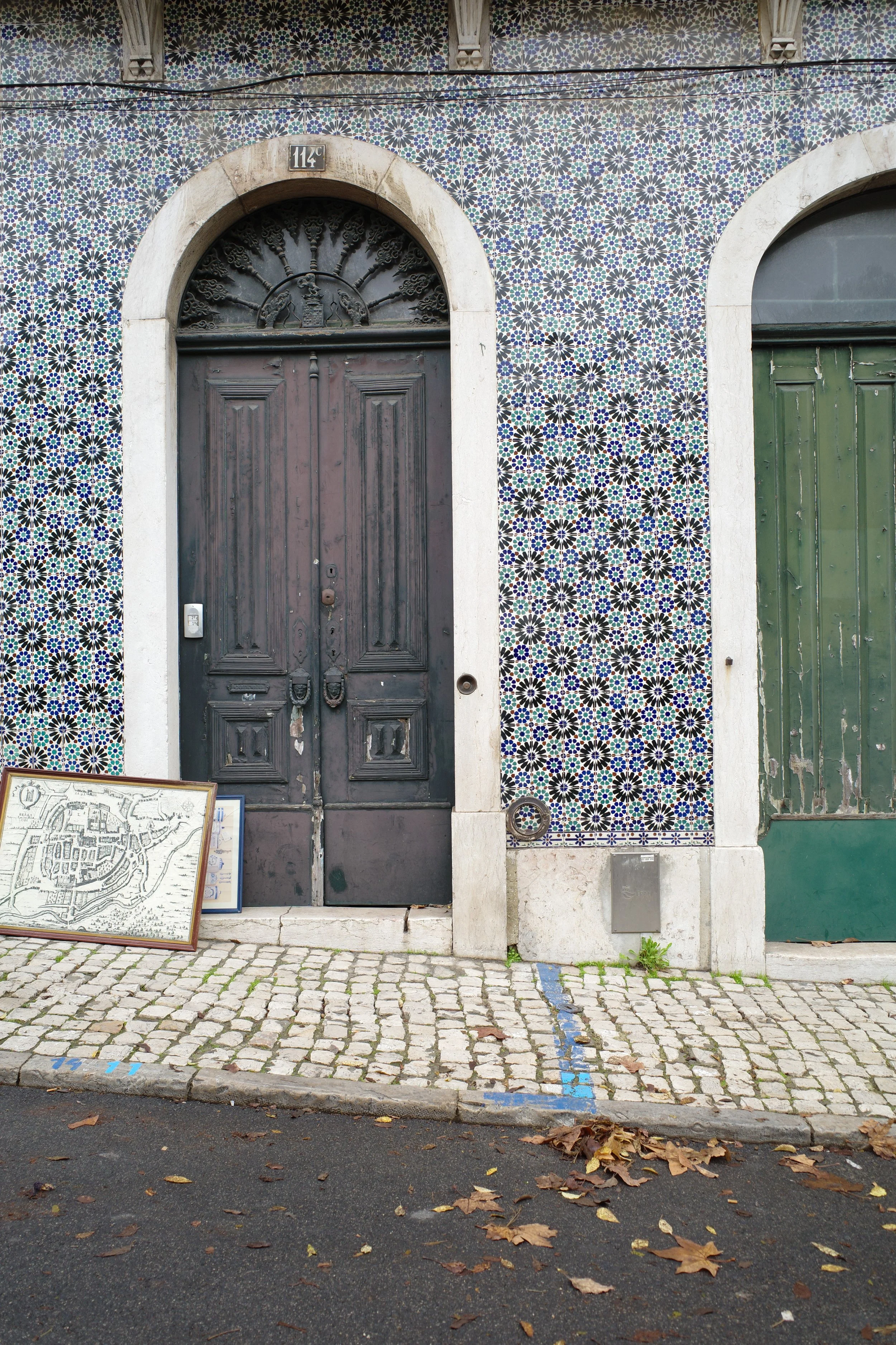 A building with ornate tile work behind a painted black wooden door with iron decorative elements. To the right, a green shed door with peeling paint. The ground is cobblestone with fallen leaves, and there are maps leaning against the wall on the si
