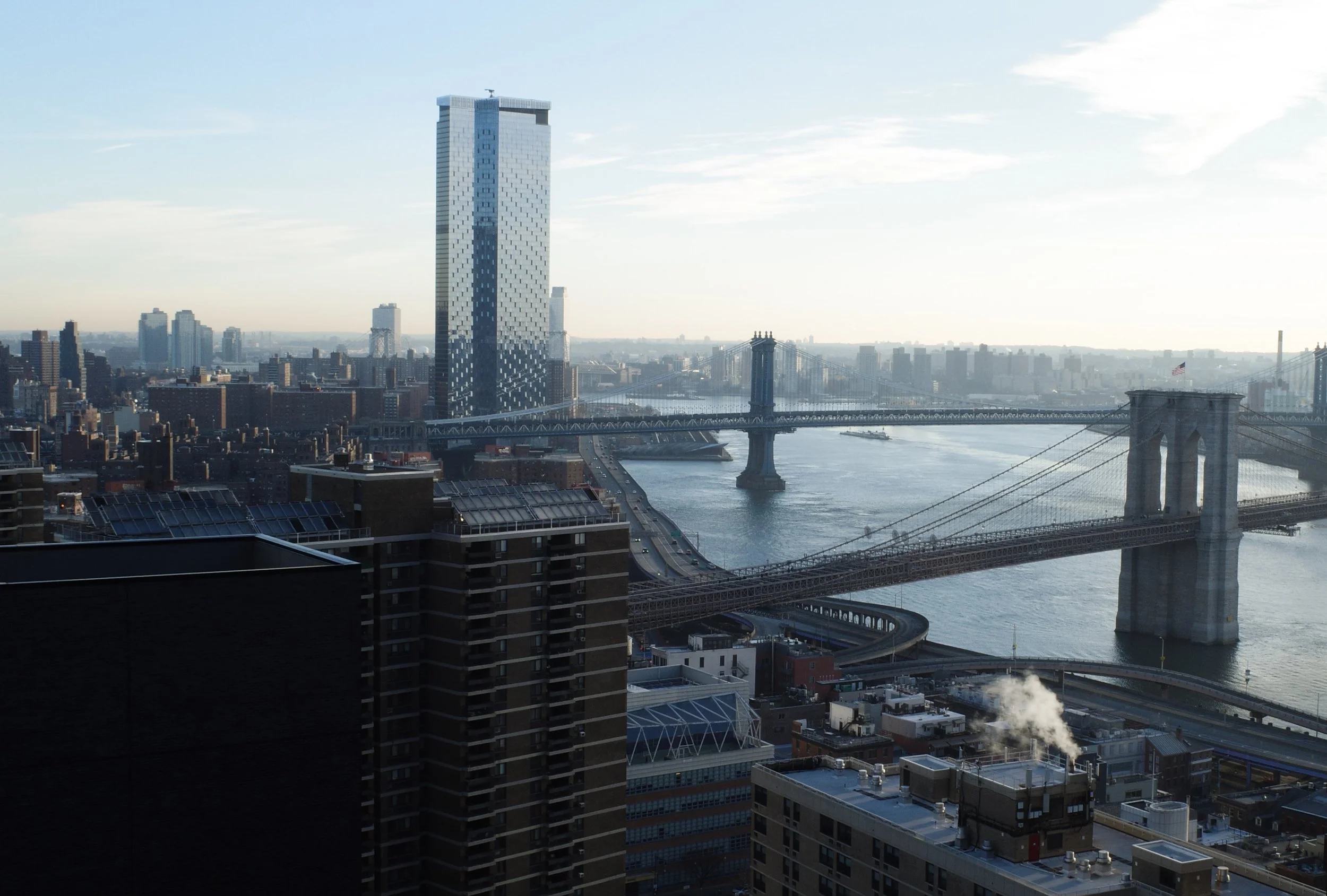City skyline featuring a tall modern glass skyscraper, Brooklyn Bridge over a river, and other city buildings with a clear sky in the background.