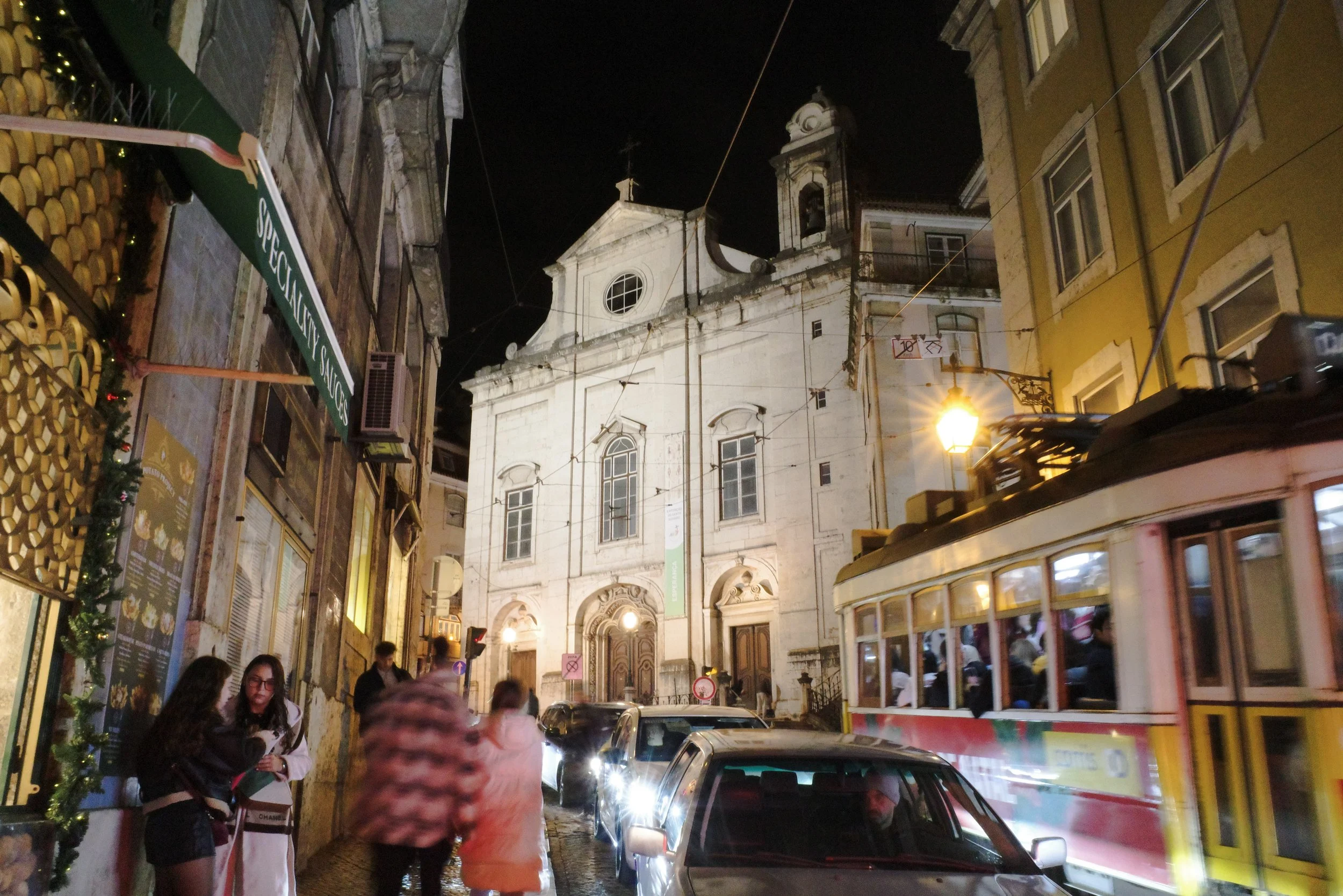 Nighttime city street scene with historic buildings, a church, and a yellow tram, with pedestrians and cars, illuminated by street lights.