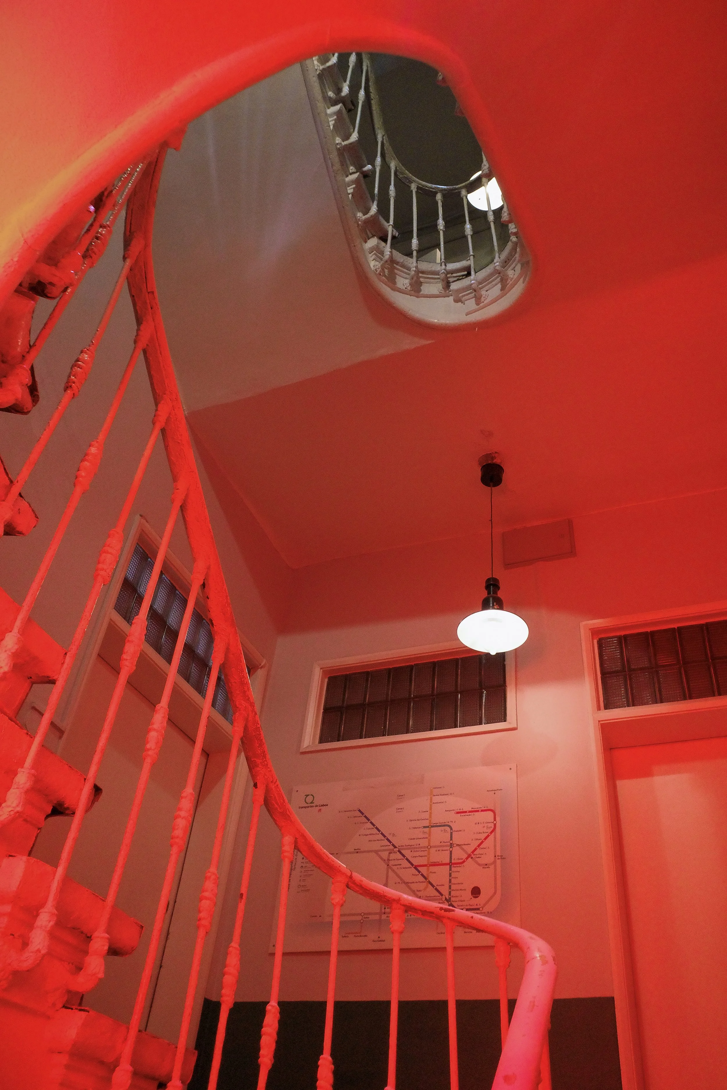 Looking up at a spiral staircase with white railings and a dark center, viewed from the ground floor, illuminated by a hanging round light fixture, with windows and a map on the wall in the background, all bathed in red lighting.